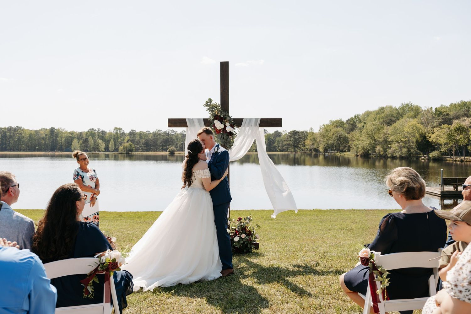 Outdoor wedding ceremony with bride and groom at floral cross altar by lake, guests seated on white chairs in sunny garden.