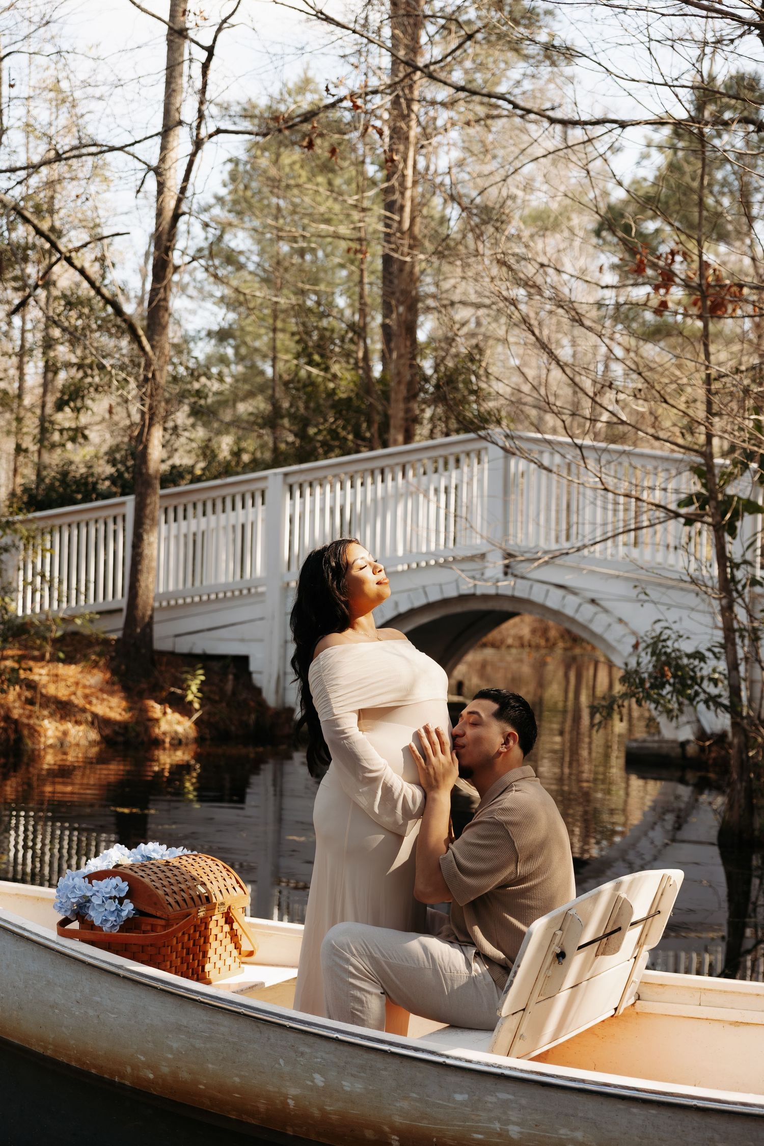 Couple in a canoe near a white bridge, woman in cream dress with partner kneeling, maternity photo shoot outdoors.
