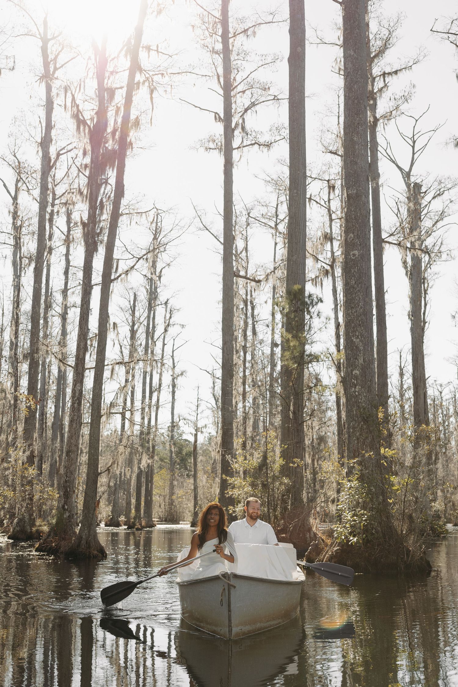 Couple in white attire rowing a boat through a misty cypress swamp forest.