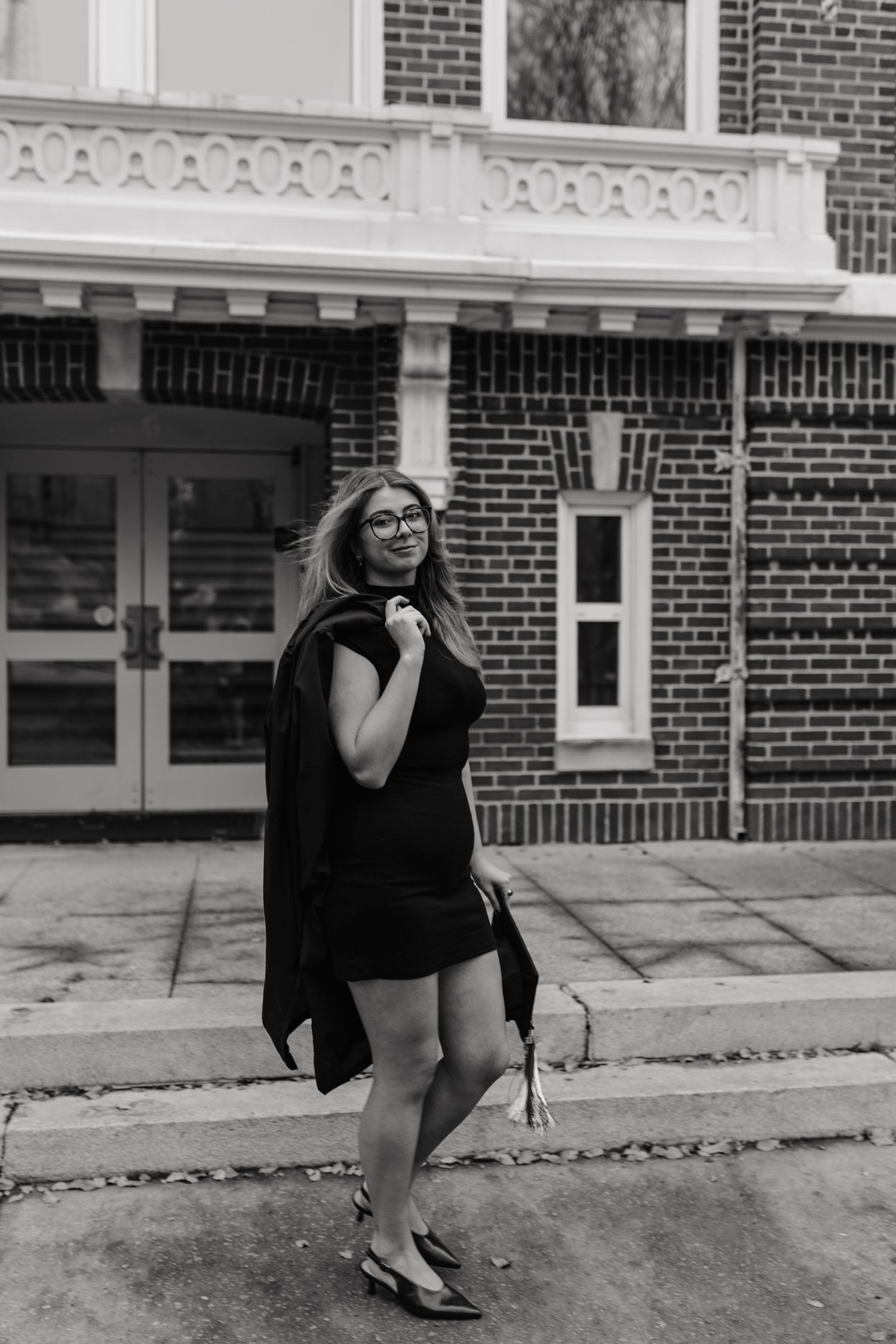 Person in black graduation gown poses playfully outside brick building in black and white photo series.