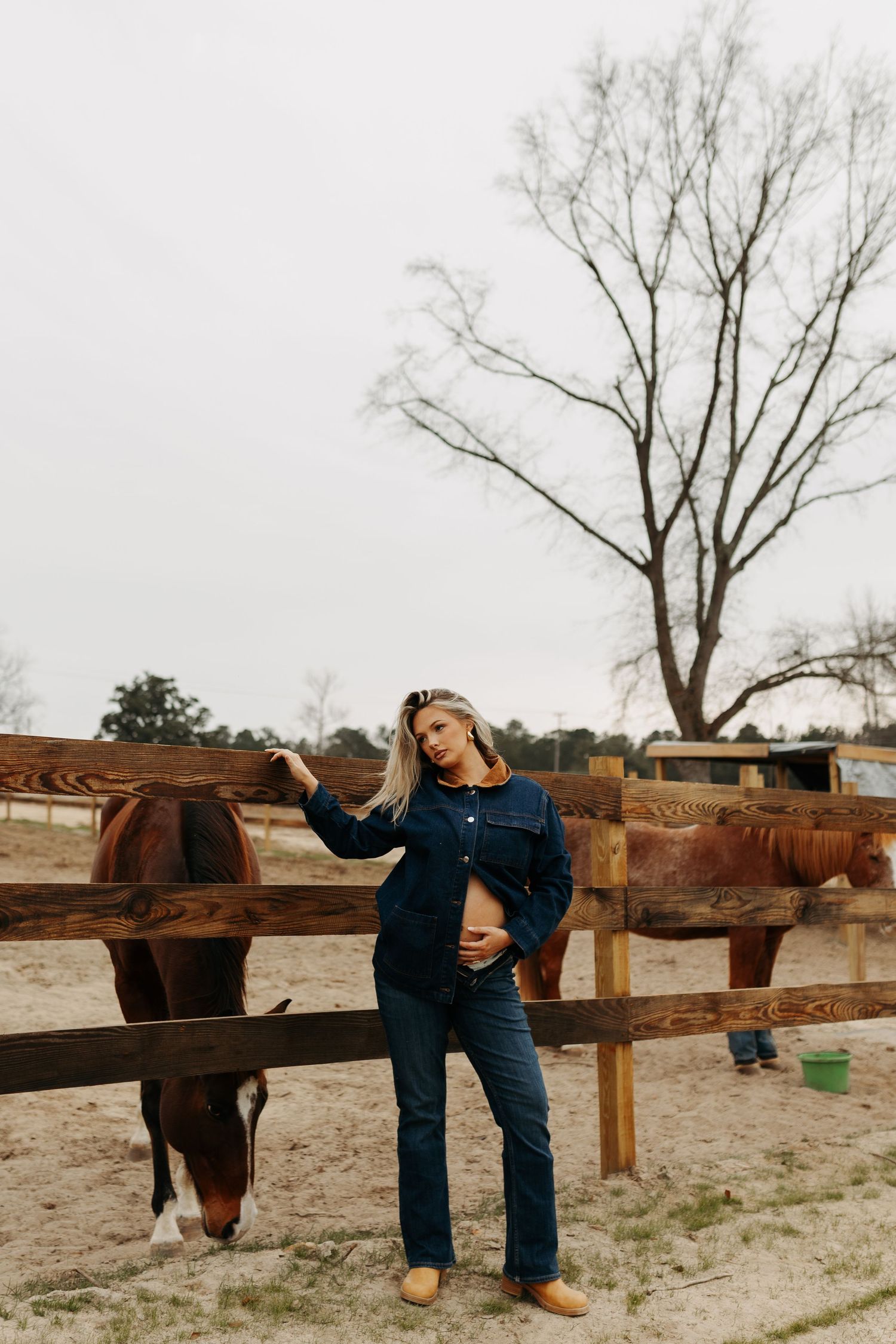 Woman in denim outfit posing by a wooden fence with a brown horse on a farm with bare trees.