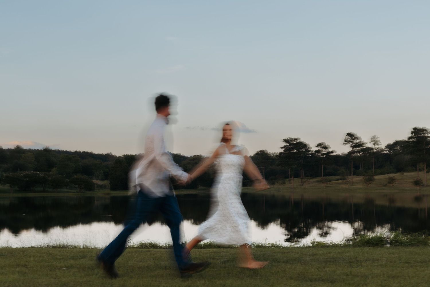 Blurred motion photo of a couple holding hands and running by a serene lake at dusk in a lush green field.