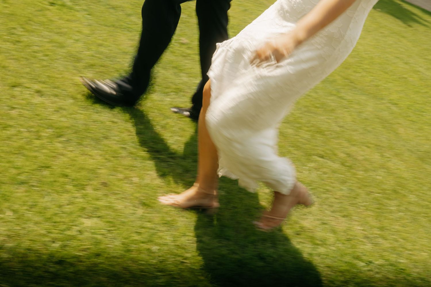Bride and groom running barefoot on green grass, white wedding dress flowing in motion blur.