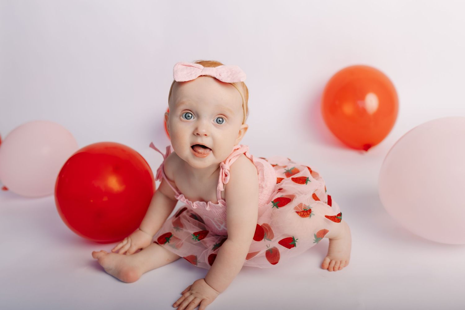 Baby girl in pink heart dress and bow headband crawls among red and pink balloons on white background.