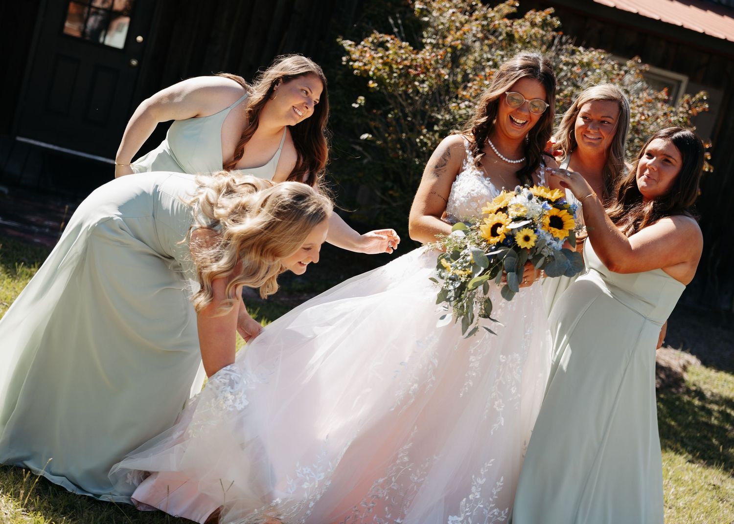 Bride in white gown laughs with bridesmaids in sage green dresses, holding sunflower bouquet outdoors.