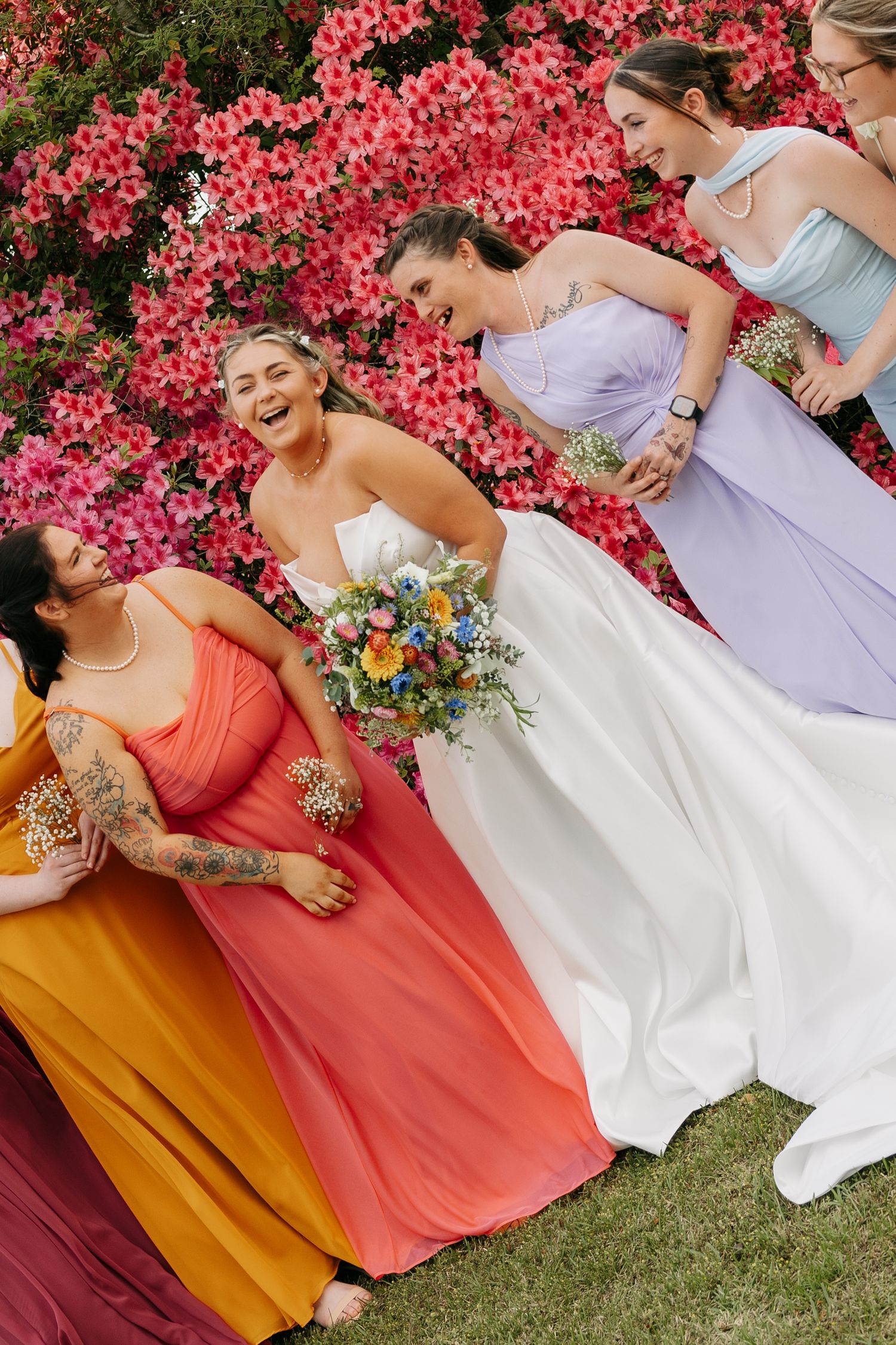 Bride laughing with colorful bridesmaids in rainbow dresses before a vibrant pink floral backdrop.