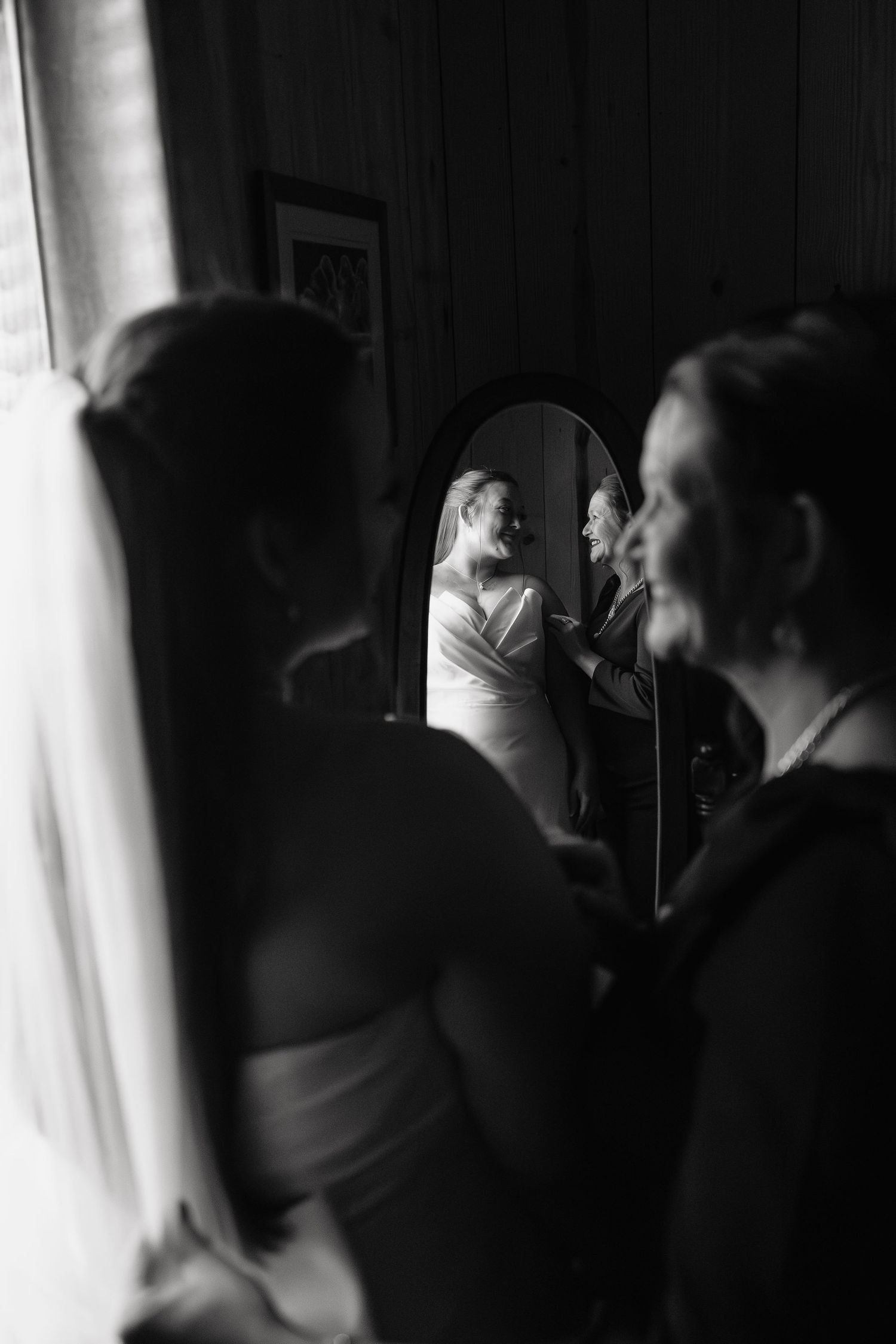 Black and white photo of bride in wedding dress reflected in mirror while bridesmaids assist her in getting ready.