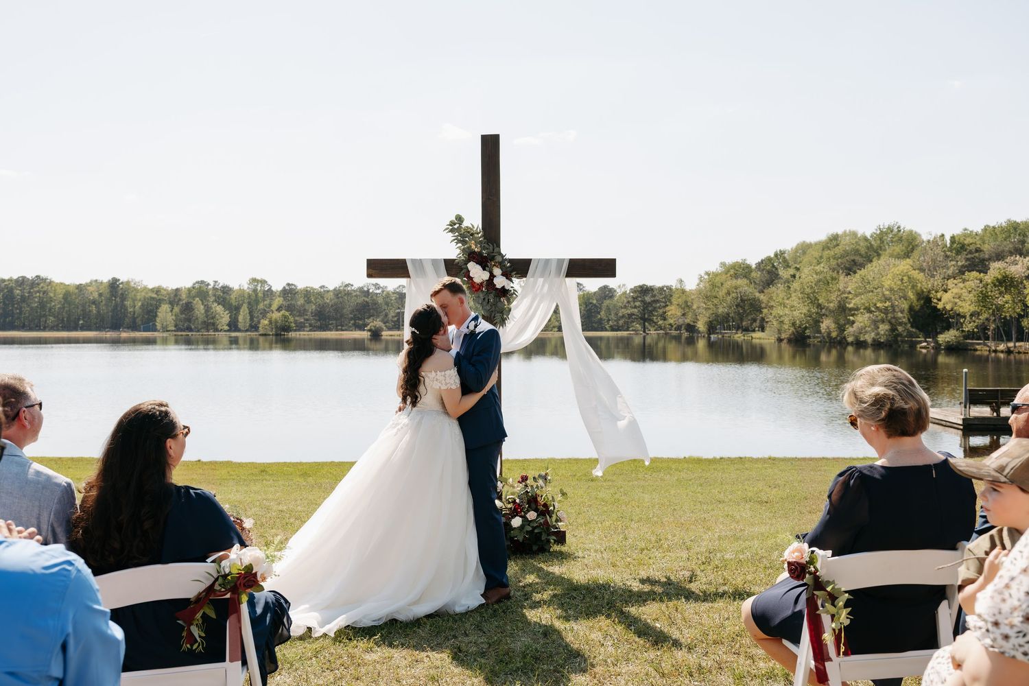 Bride and groom exchange vows at outdoor lakeside wedding ceremony with wooden cross altar and seated guests.