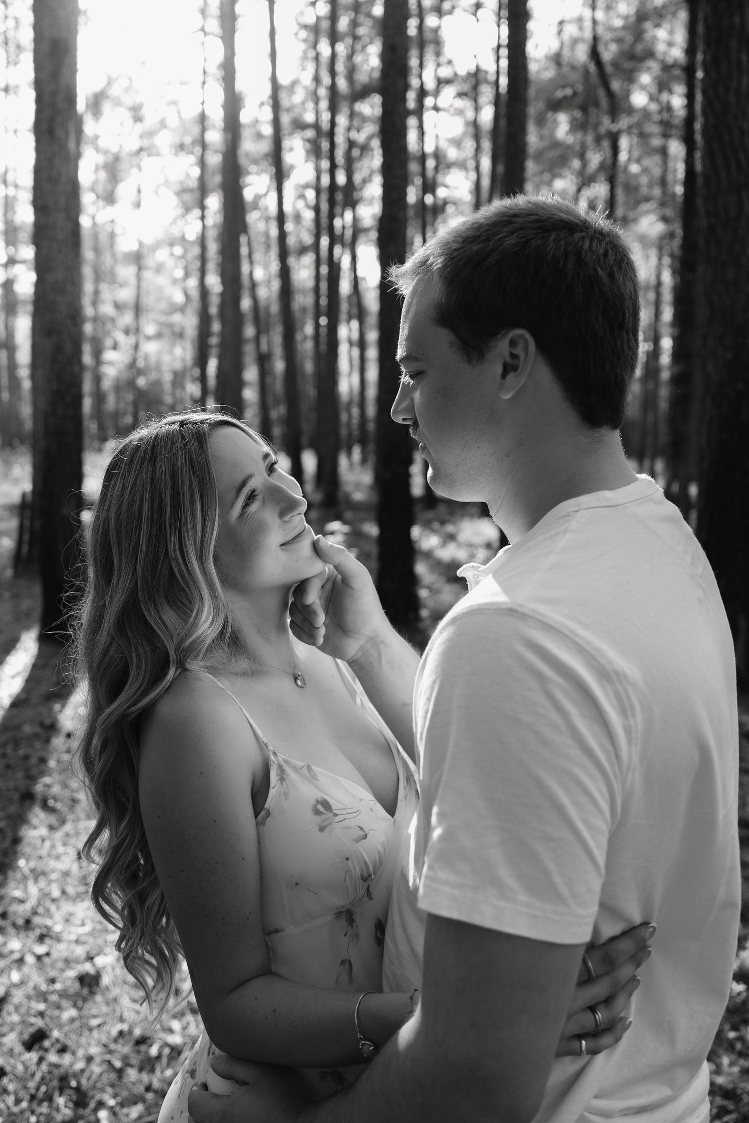 Black and white photo of a romantic couple sharing a tender moment in a sunlit forest, man gently touching woman's face.