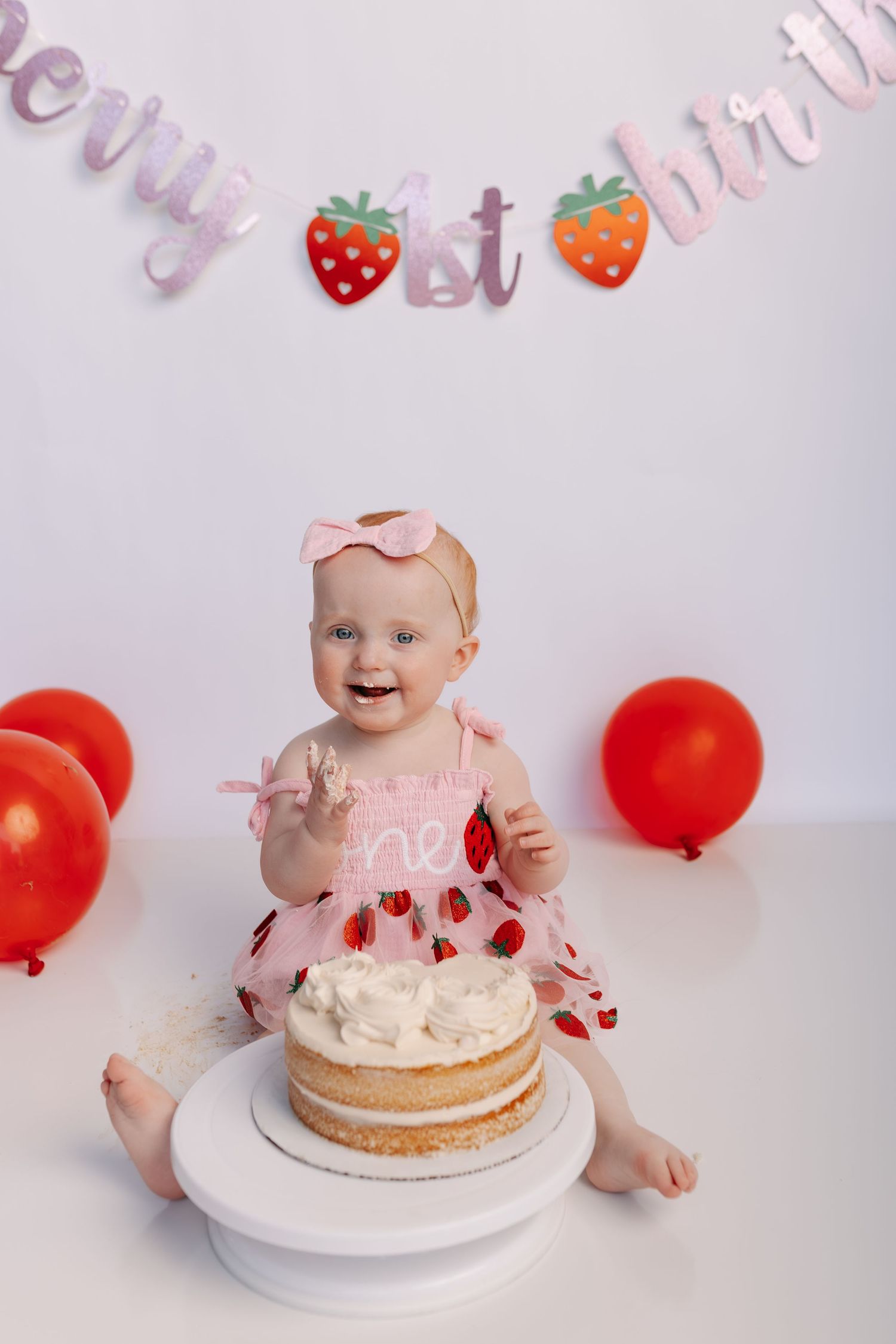 Baby girl in strawberry dress smashes her first birthday cake surrounded by red balloons and a berry 1st birthday banner.