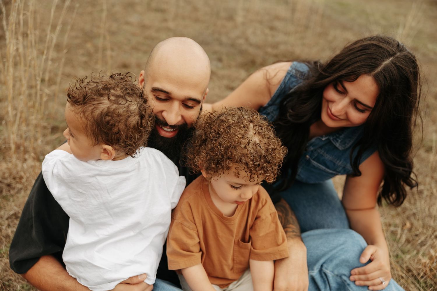 Happy family with two curly-haired children sitting together outdoors in a golden field, sharing a joyful moment.