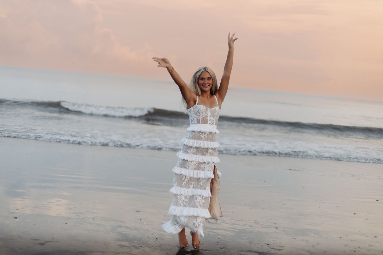 Woman in white lace dress celebrating on beach at sunset with arms raised.
