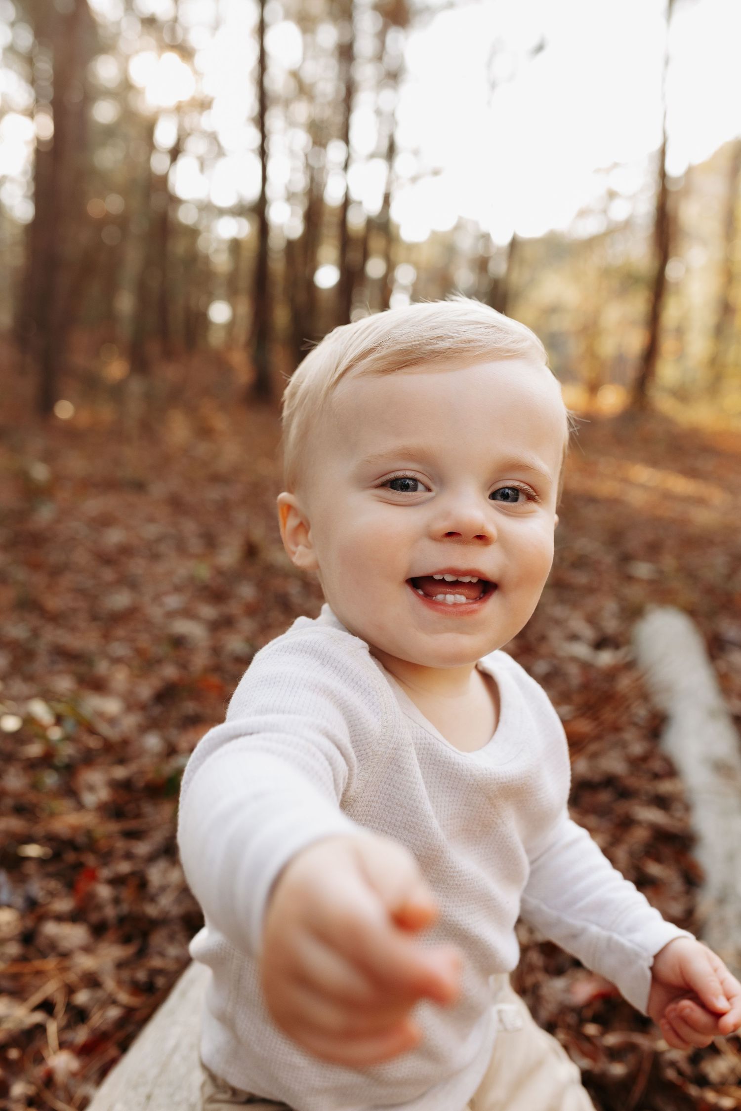 Smiling toddler in white shirt reaching toward camera in sunlit autumn forest with fallen leaves.