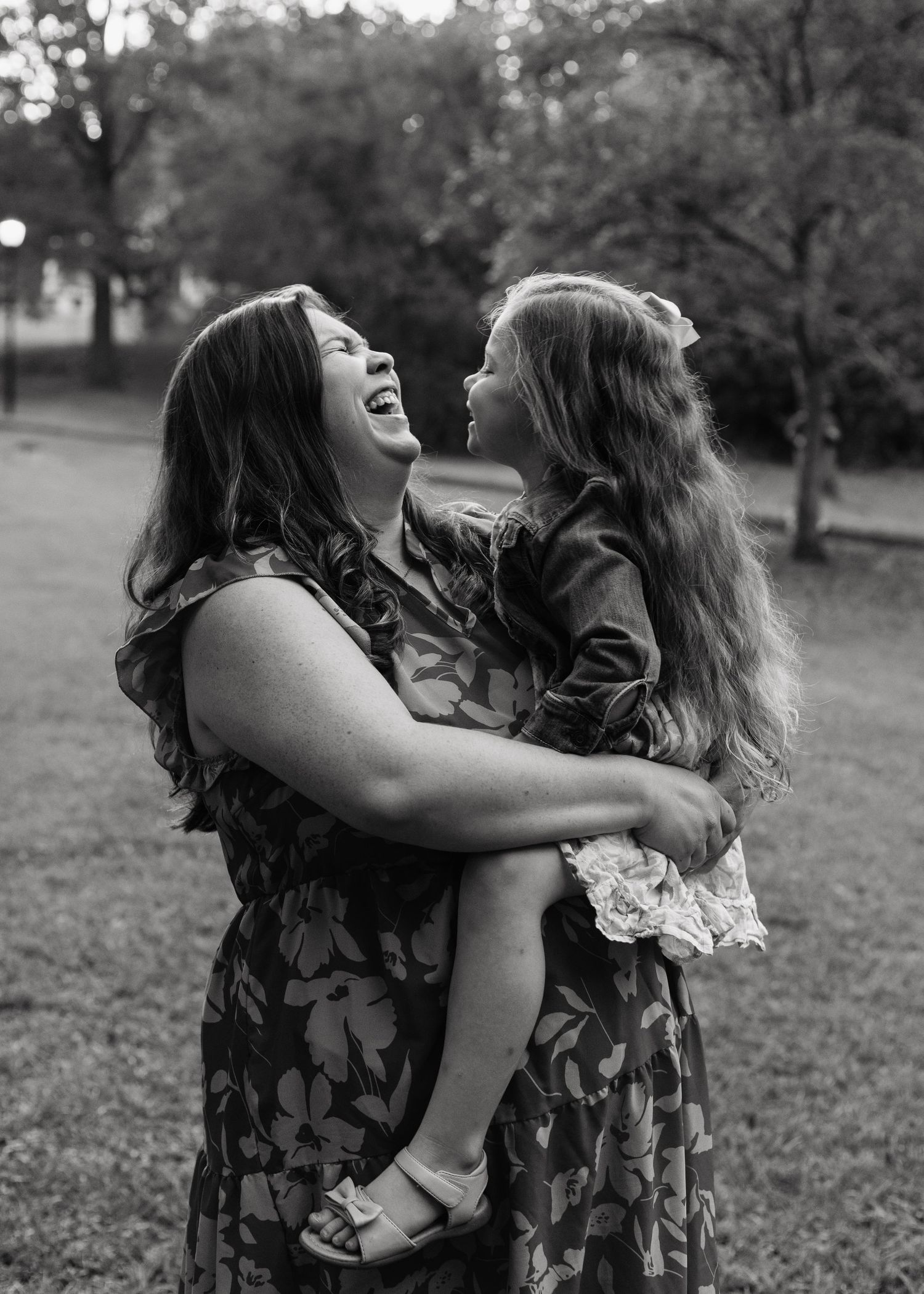 Black and white photo of a laughing mother holding her young daughter outdoors in a park setting.