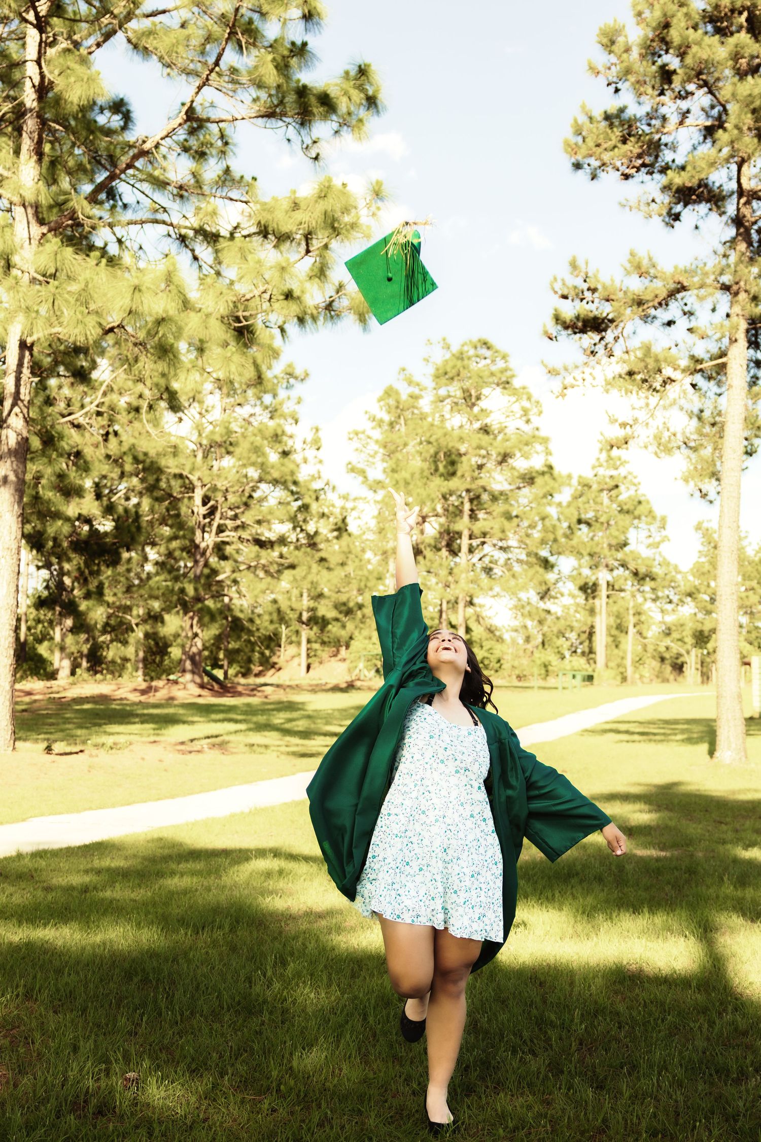 Graduate in green cap and gown twirls joyfully in a sunlit field surrounded by pine trees.