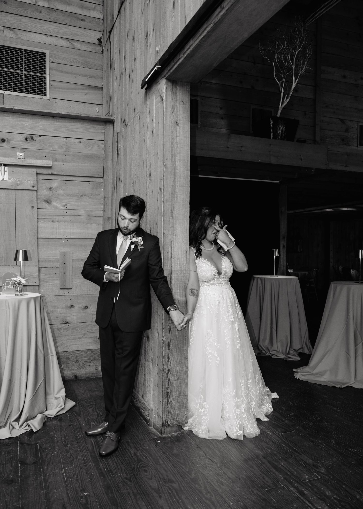 Black and white photo of bride and groom holding hands, reading vows against a rustic barn wooden pillar.