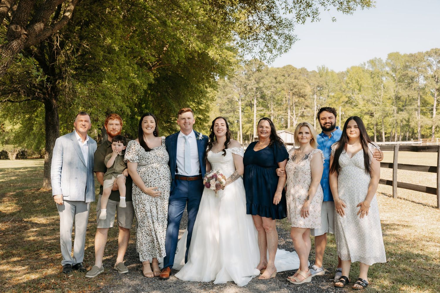 Wedding party group photo outdoors with bride and groom surrounded by family and friends near a wooden fence.