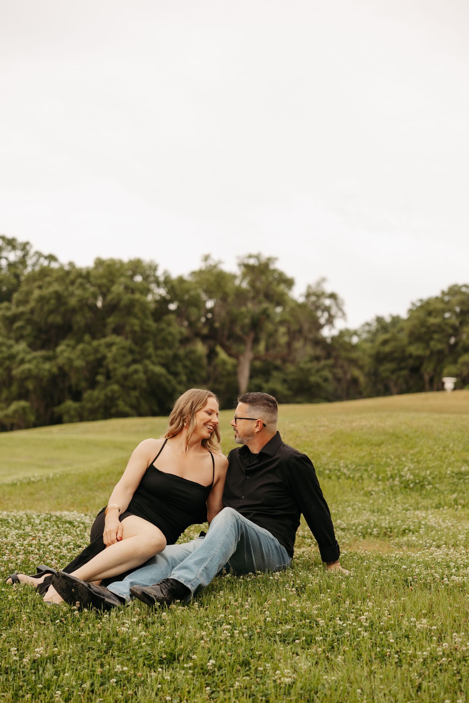 Couple sitting together on green grass field, woman in black dress and man in black shirt and jeans, smiling at each other.