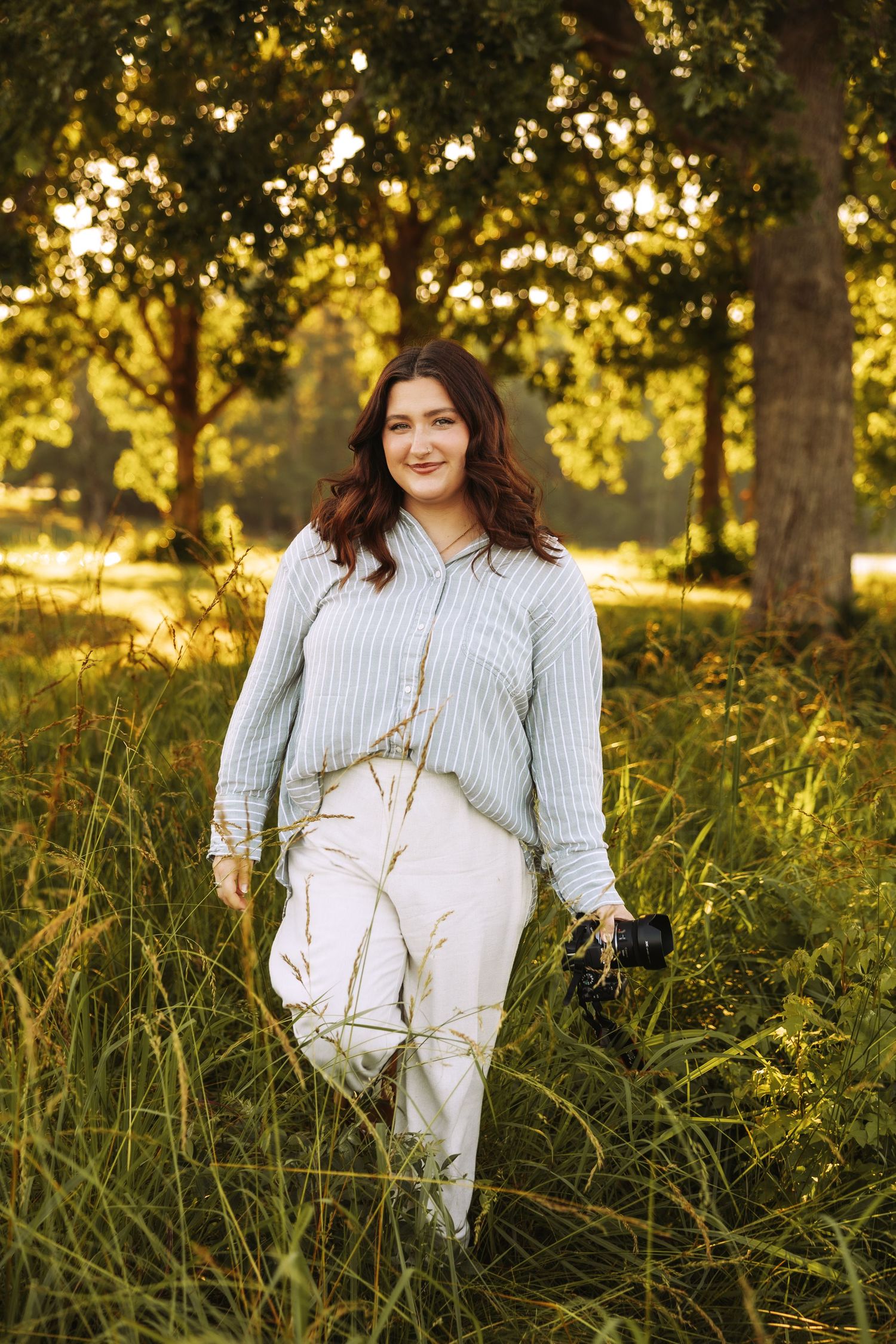 Woman in striped shirt and white pants holding camera standing in lush green park during golden hour.