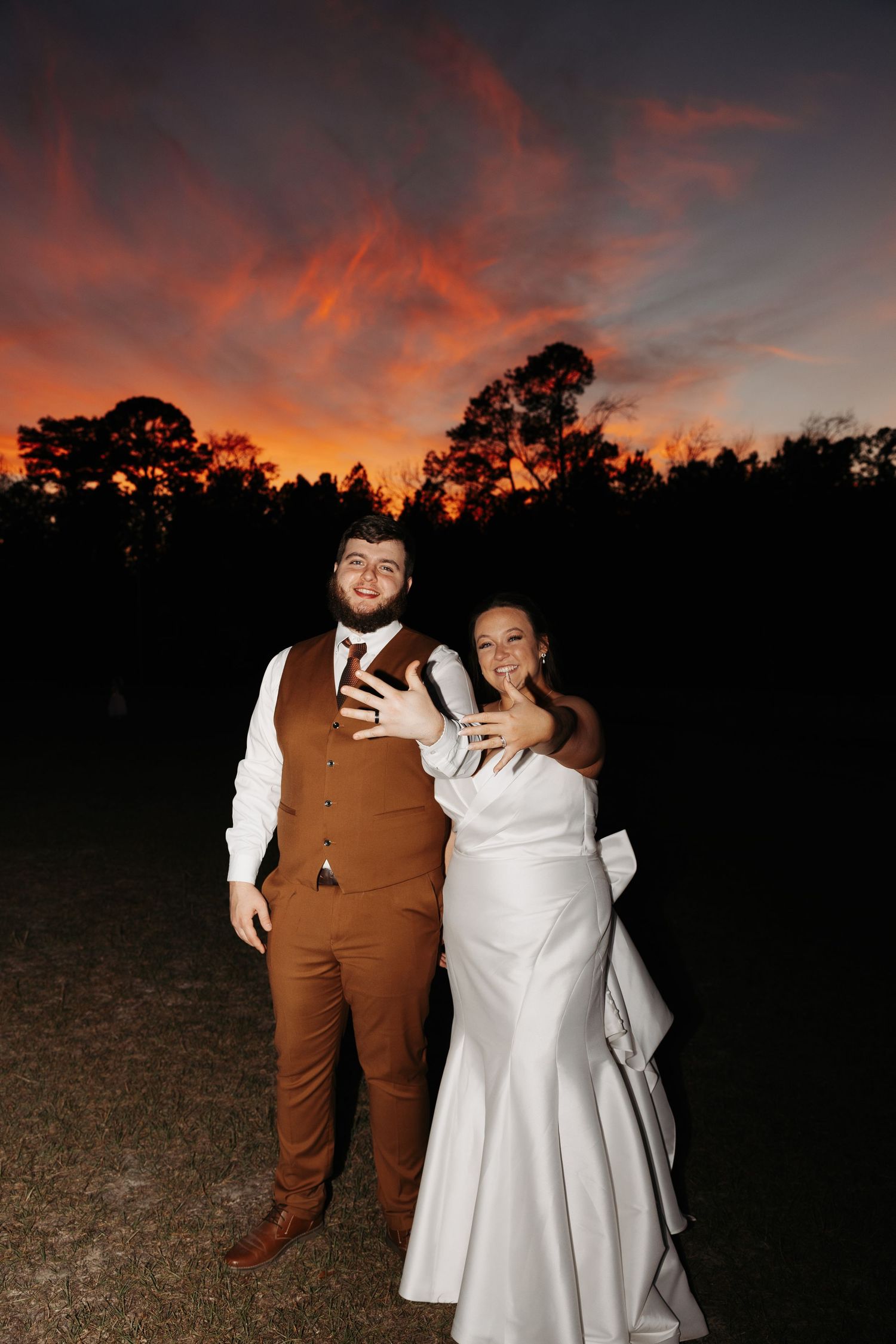 Bride and groom pose together showing rings against a dramatic red and orange sunset sky.