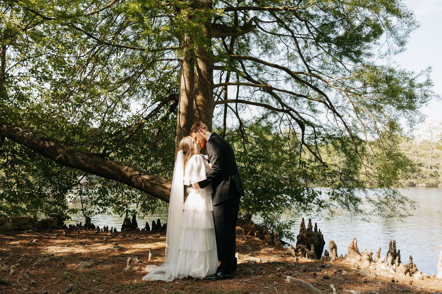 Bride and groom kissing under a large tree by a lake, surrounded by lush greenery on their wedding day.