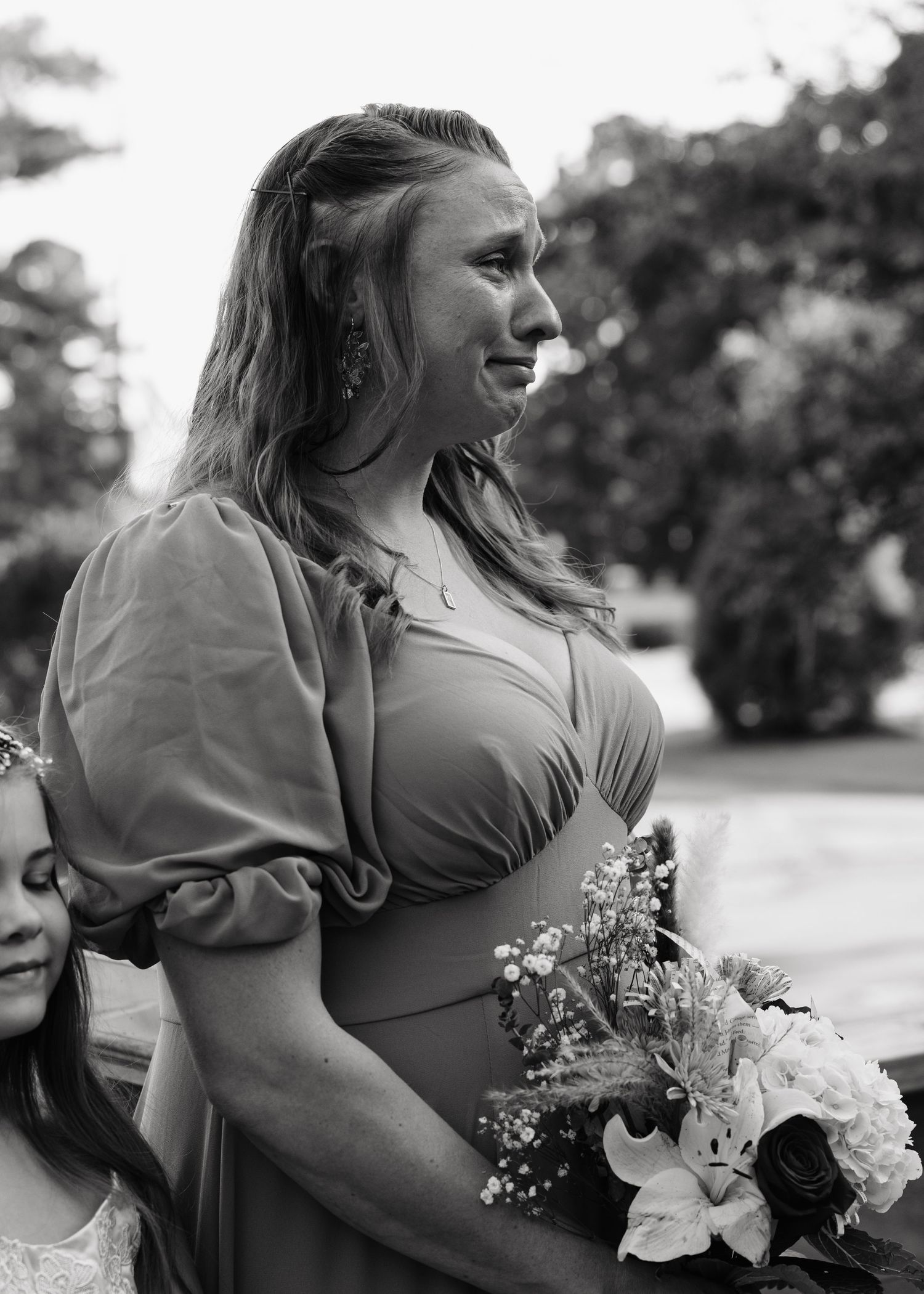 Emotional bridesmaid in sage green dress holding white floral bouquet during outdoor wedding ceremony in black and white photo.