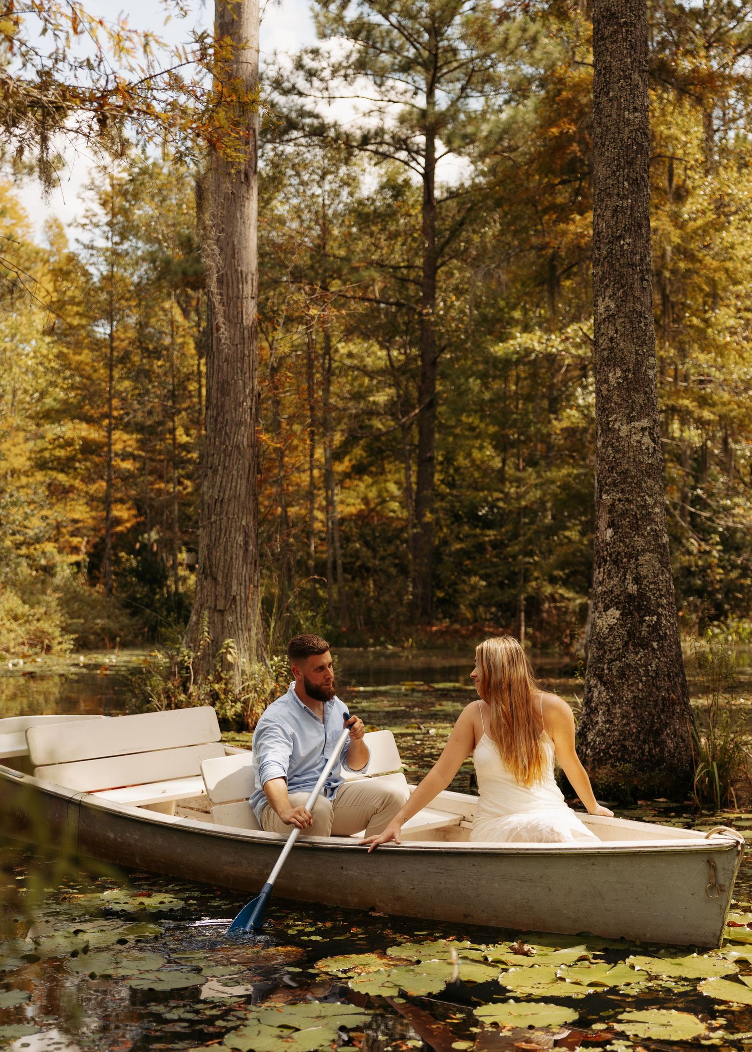 Couple holding hands in a rowboat surrounded by lily pads and autumn trees in a serene forest lake.