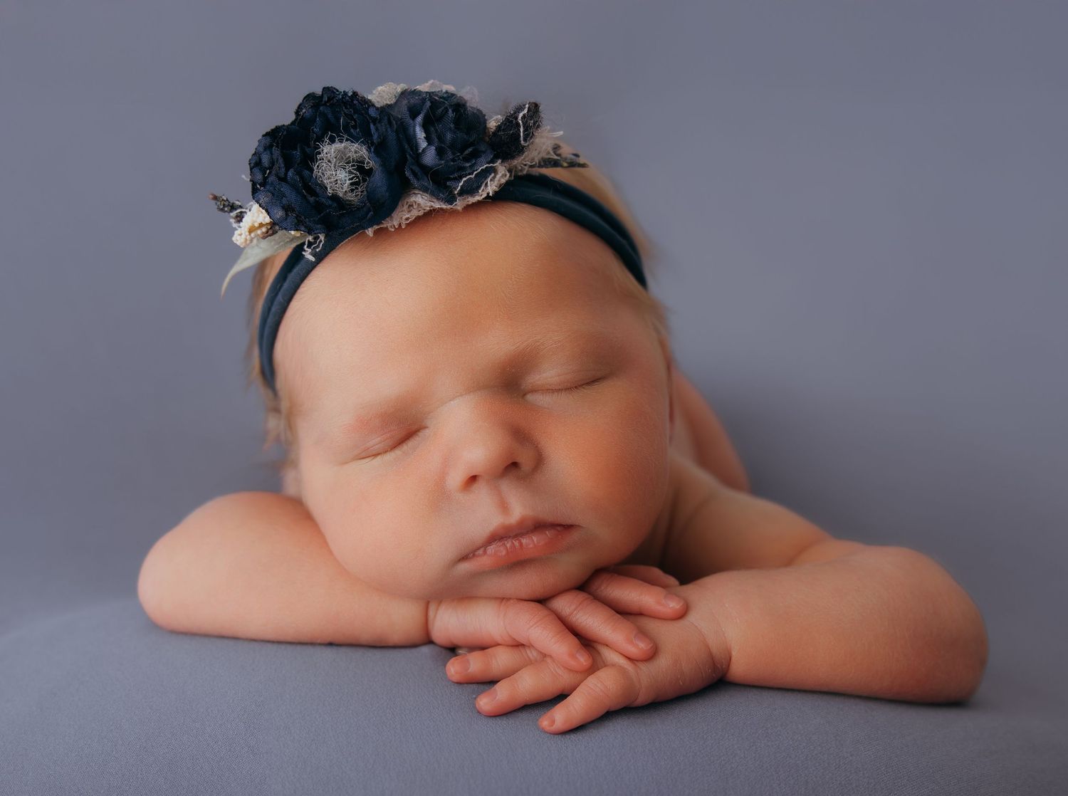 Peaceful newborn baby girl sleeping with chin on hands, wearing a navy floral headband on a gray backdrop.