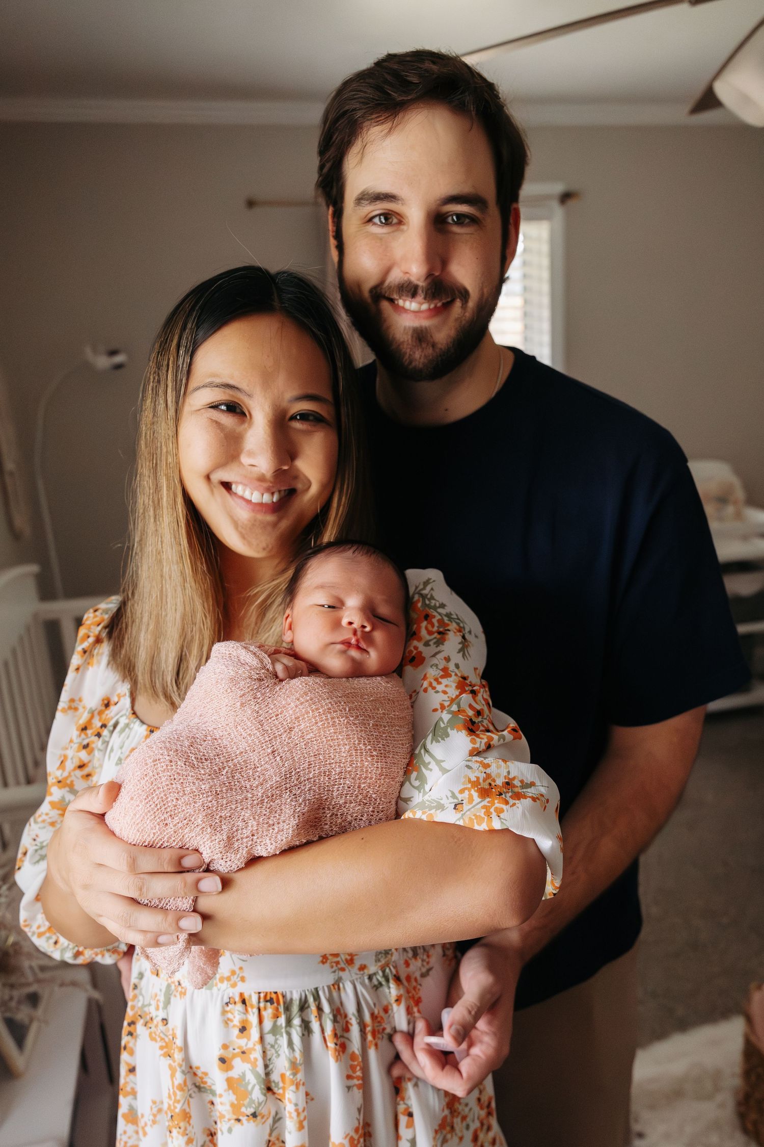 A couple stands together indoors holding their newborn baby swaddled in a pink blanket.