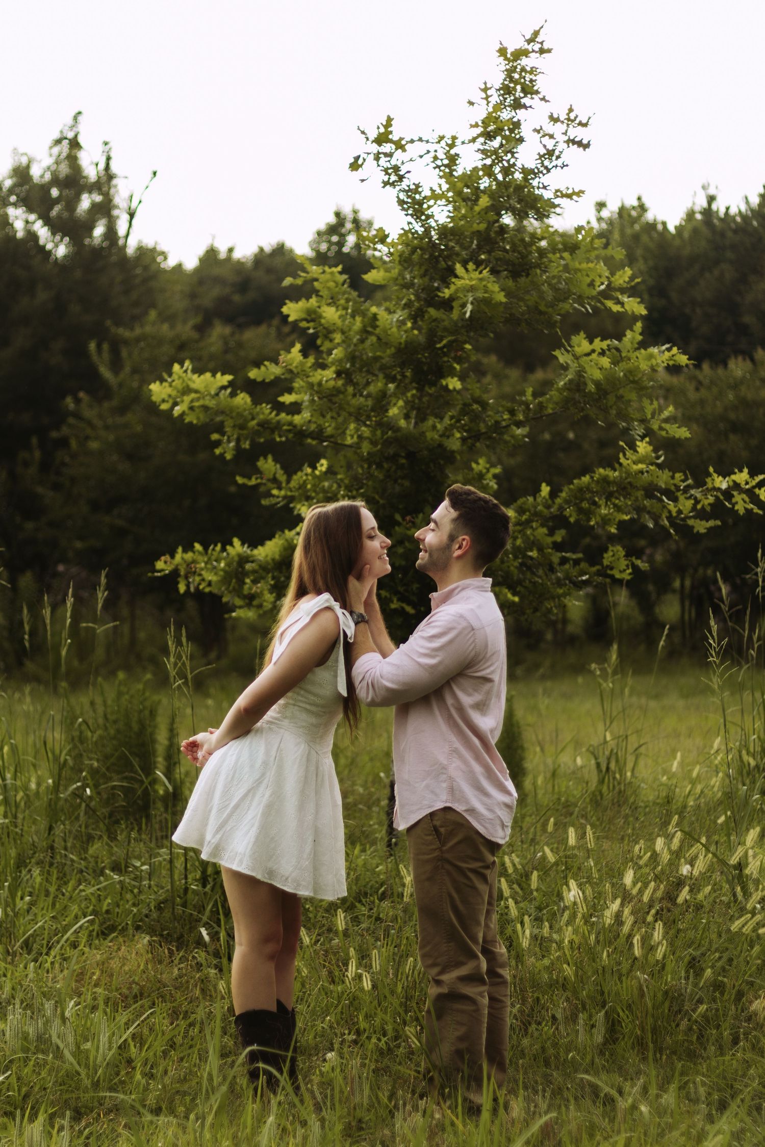 A couple in summery attire laughs together while standing in a sunlit meadow with trees in the background.