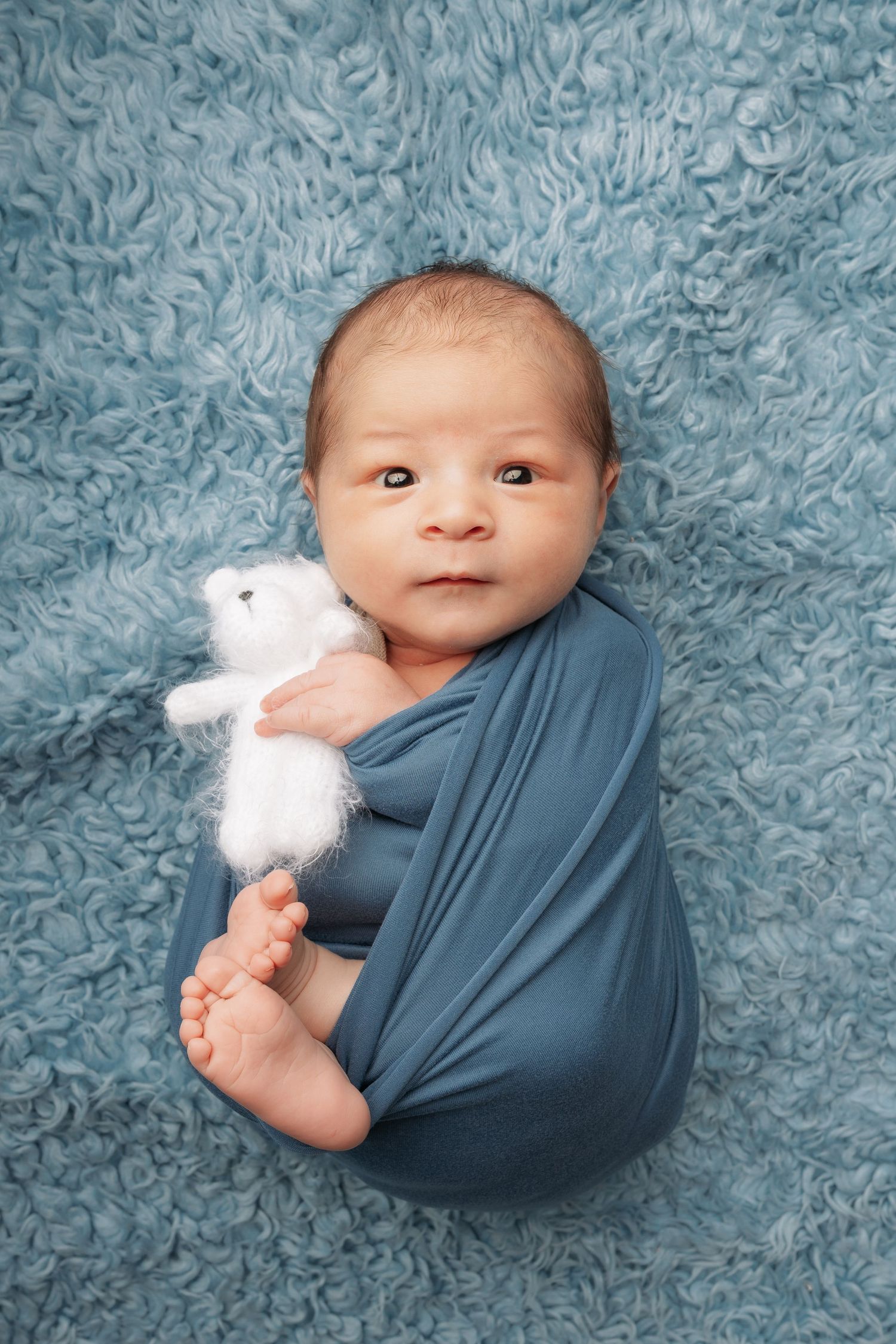 Baby wrapped in blue swaddle lying on blue textured blanket with white stuffed bunny toy in progressive poses.