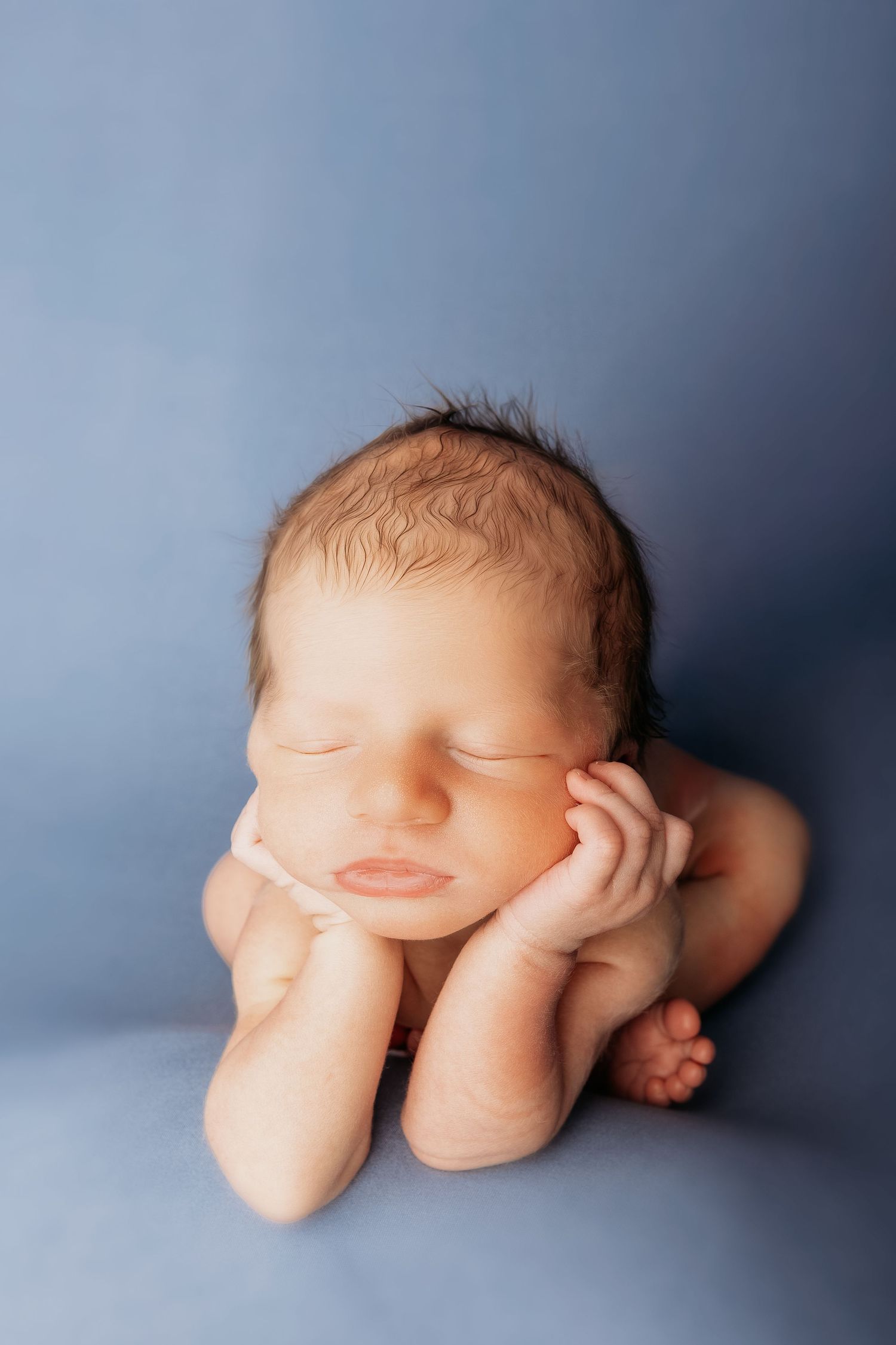 A sleeping newborn baby lies on a soft blue backdrop in a peaceful curled position.