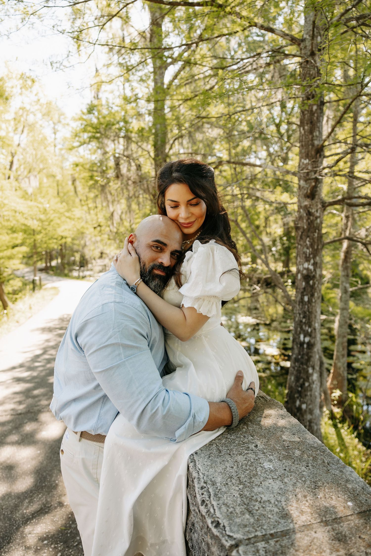 Couple embracing outdoors, pregnant woman in white dress with bald bearded man, surrounded by lush green trees.