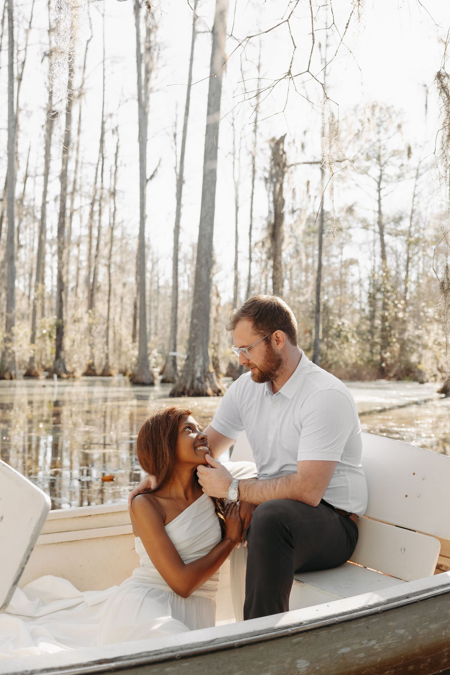 A couple in white attire shares a romantic moment on a boat surrounded by cypress trees in a tranquil swamp.