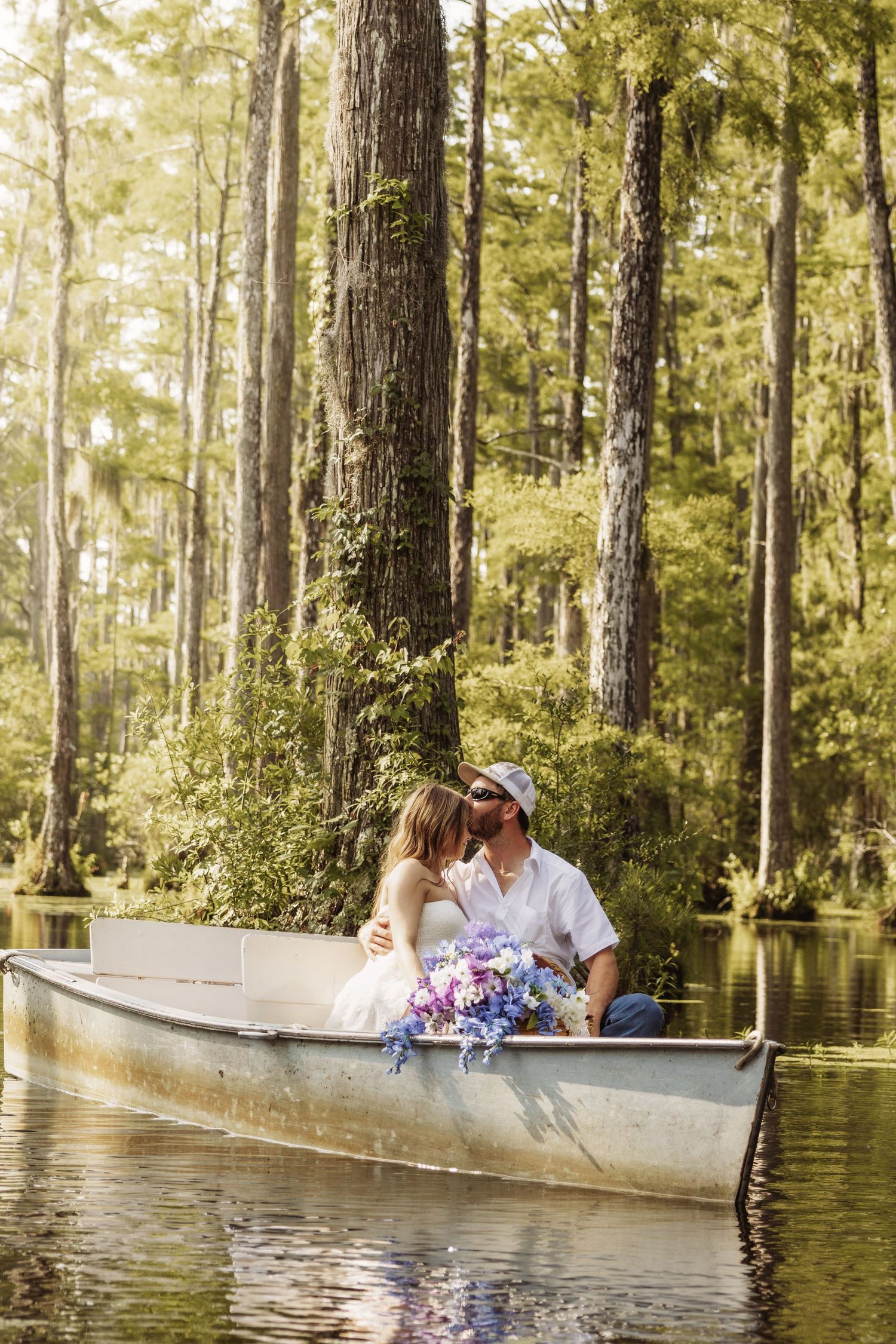 A couple shares a romantic kiss in a canoe surrounded by tall trees and lush greenery, bride holding a purple bouquet.