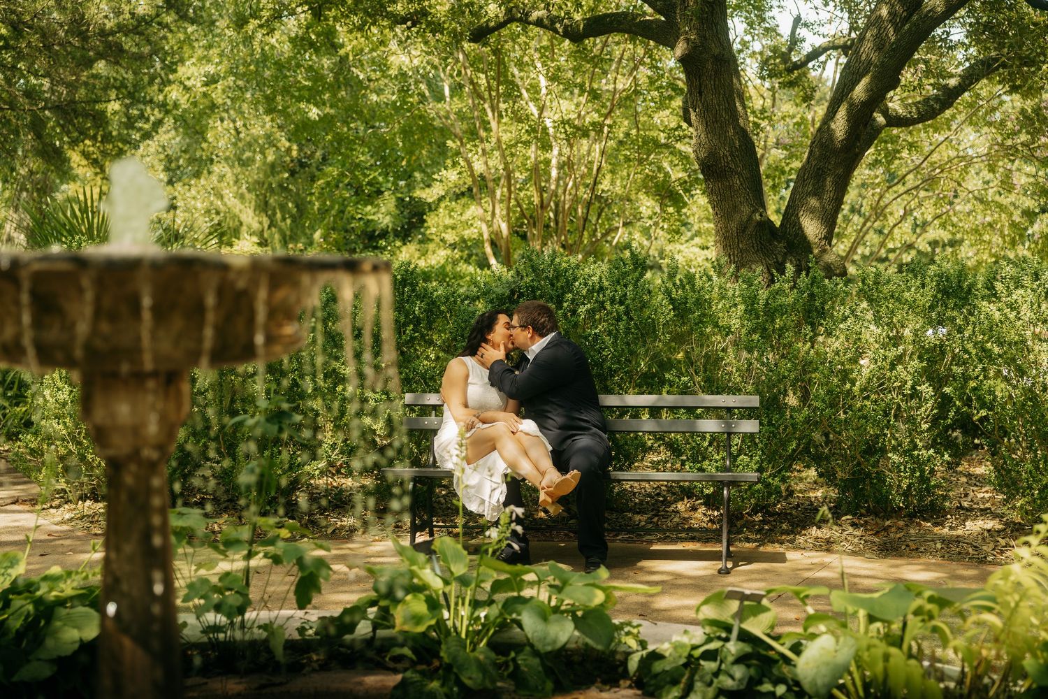 Couple sharing a romantic kiss on a bench surrounded by lush greenery in a garden setting.