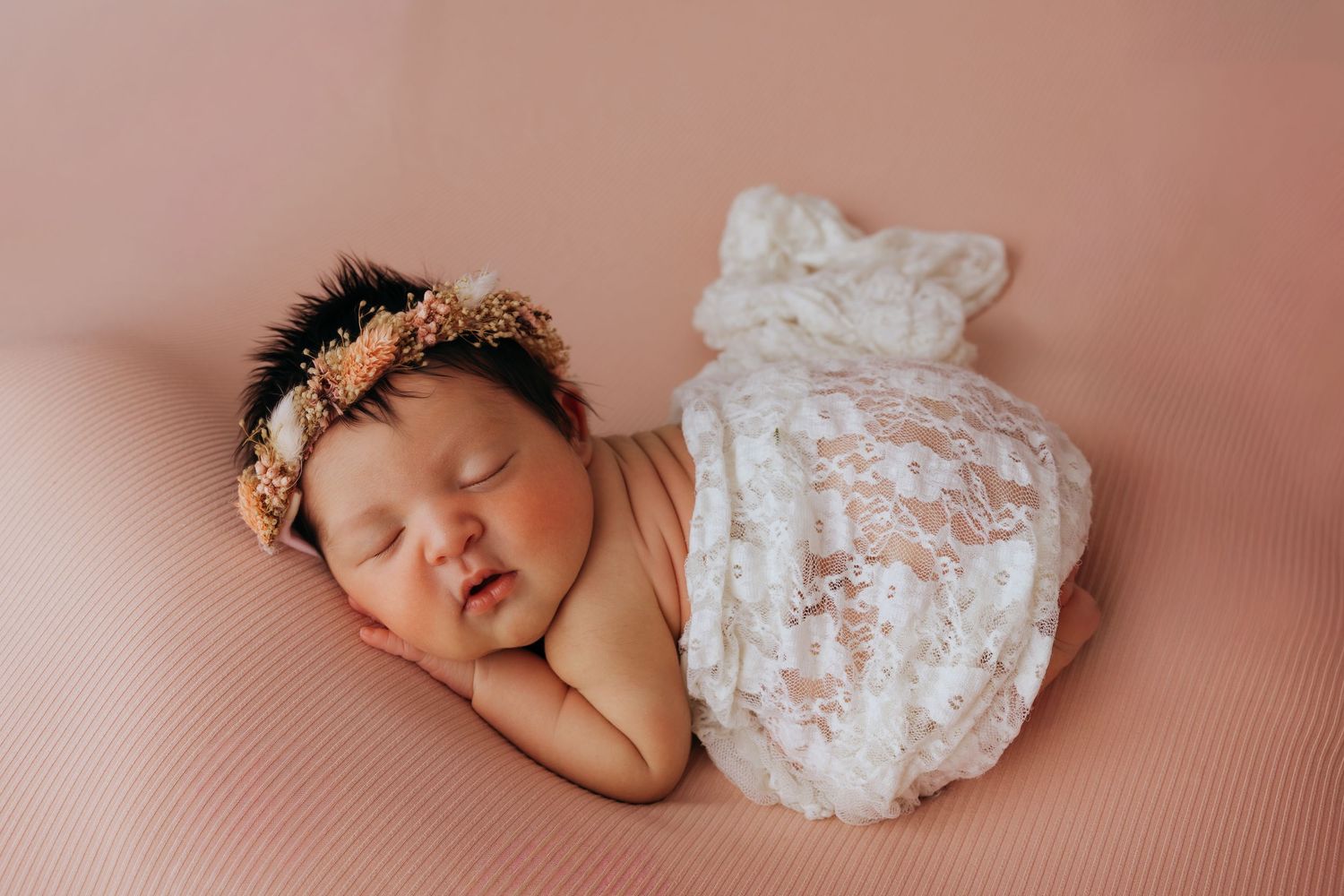 Sleeping newborn baby girl wearing a floral headband, wrapped in white lace on a pink background.