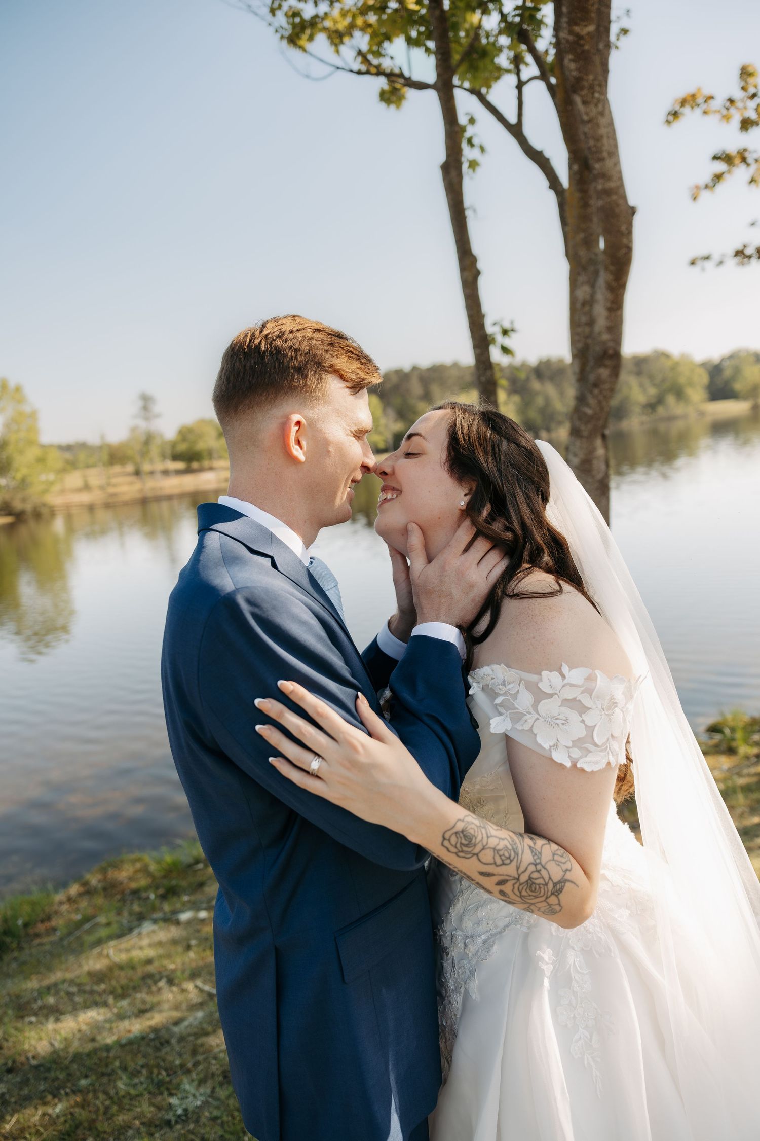 Bride and groom share a romantic moment by a sunlit lake, groom tenderly holding brides face before a kiss.