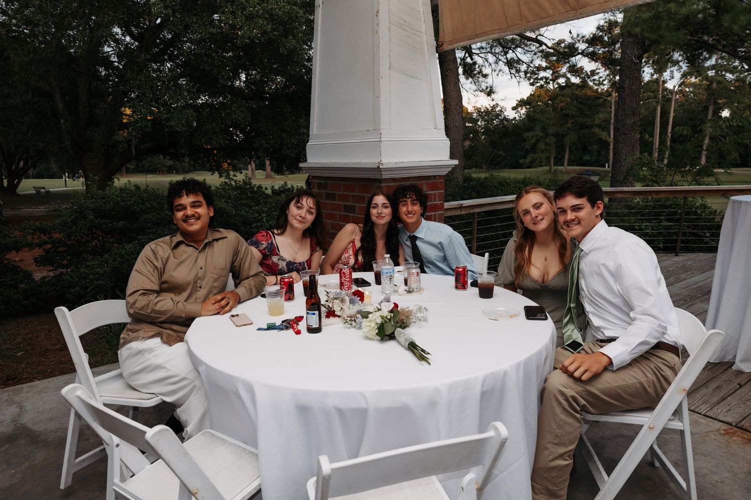 Six wedding reception guests smiling at a round table with white chairs in an outdoor venue at dusk.