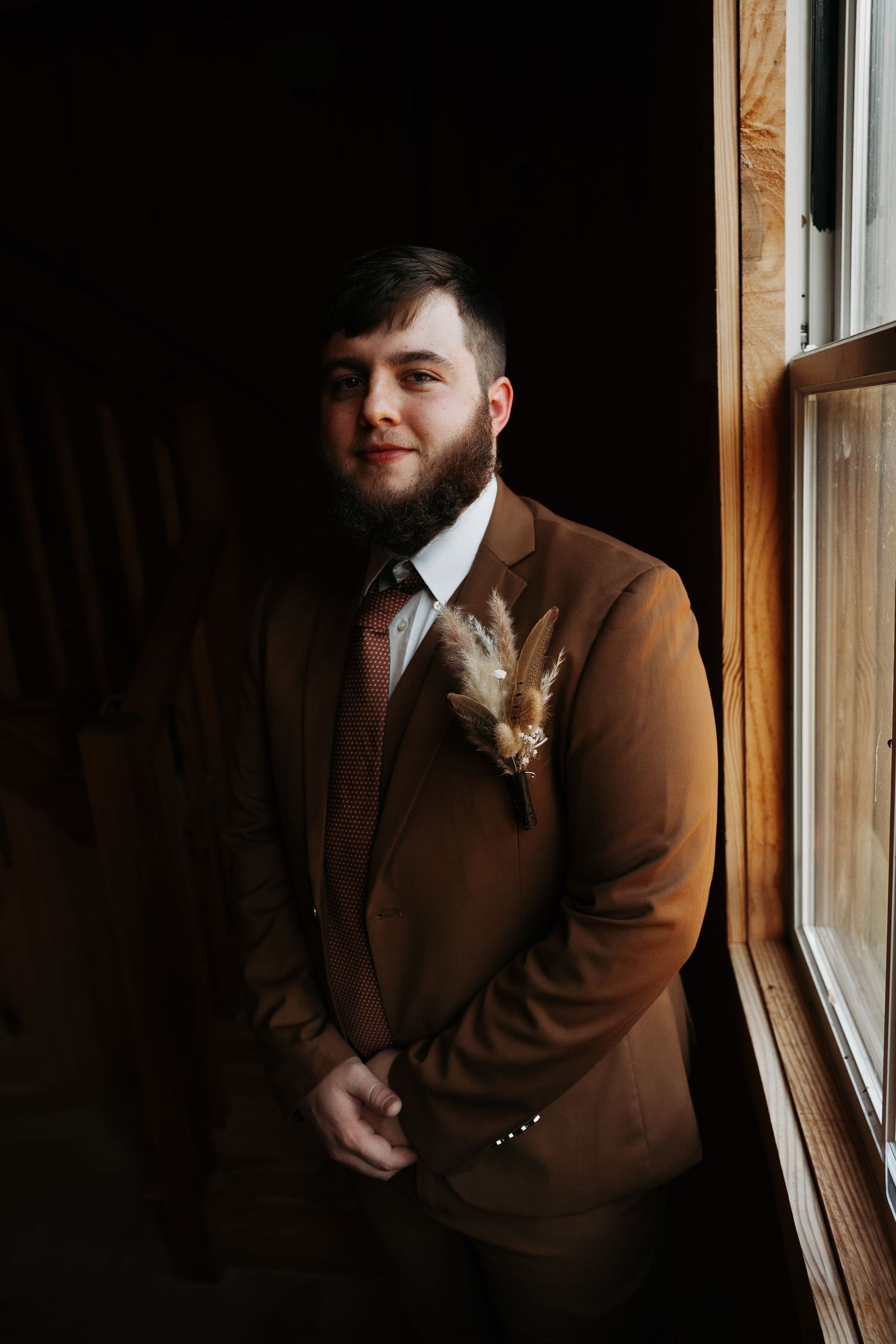 Groom in brown suit with pampas grass boutonniere posing by rustic barn window.