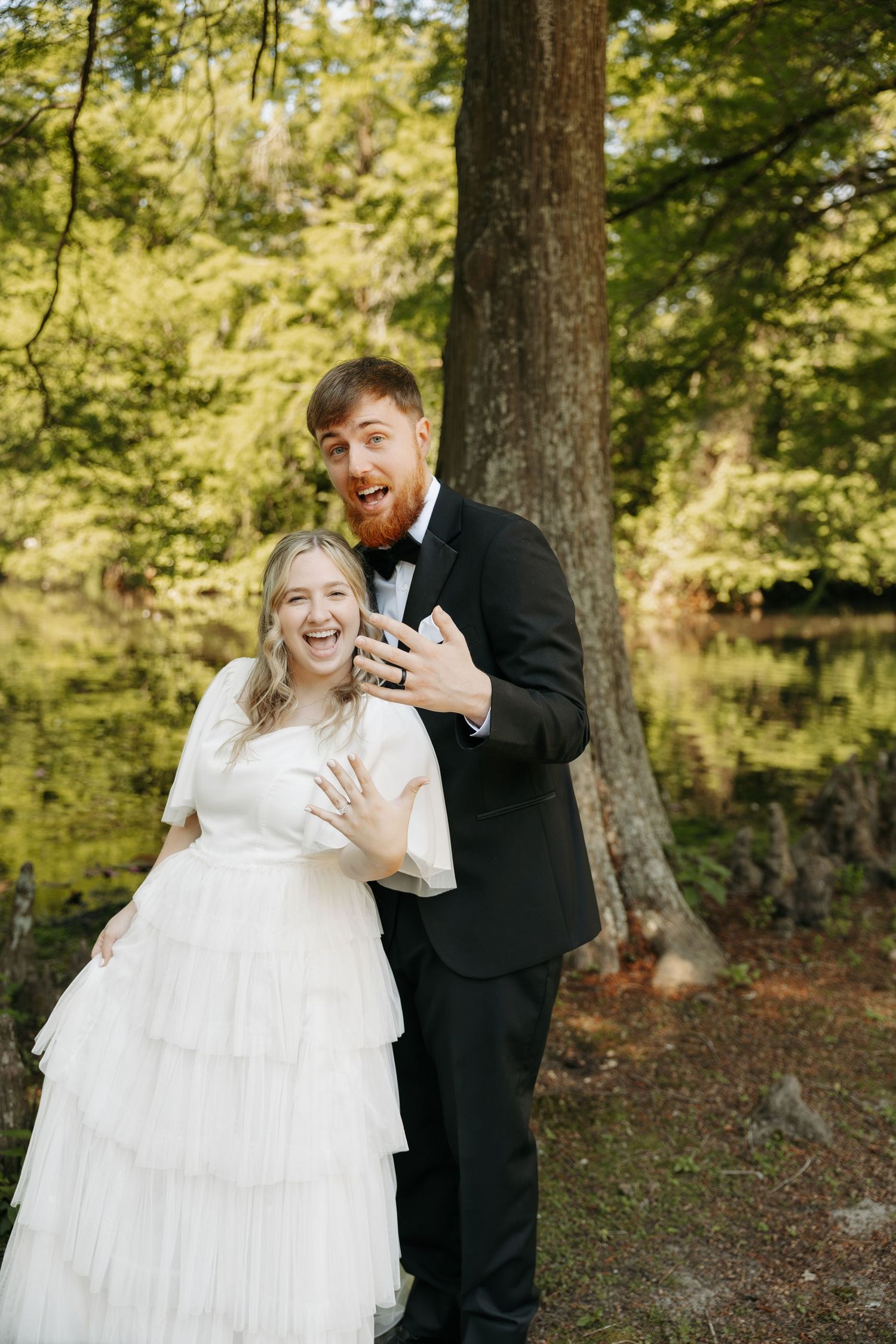 Joyful bride and groom showing wedding rings outdoors by a forest lake.