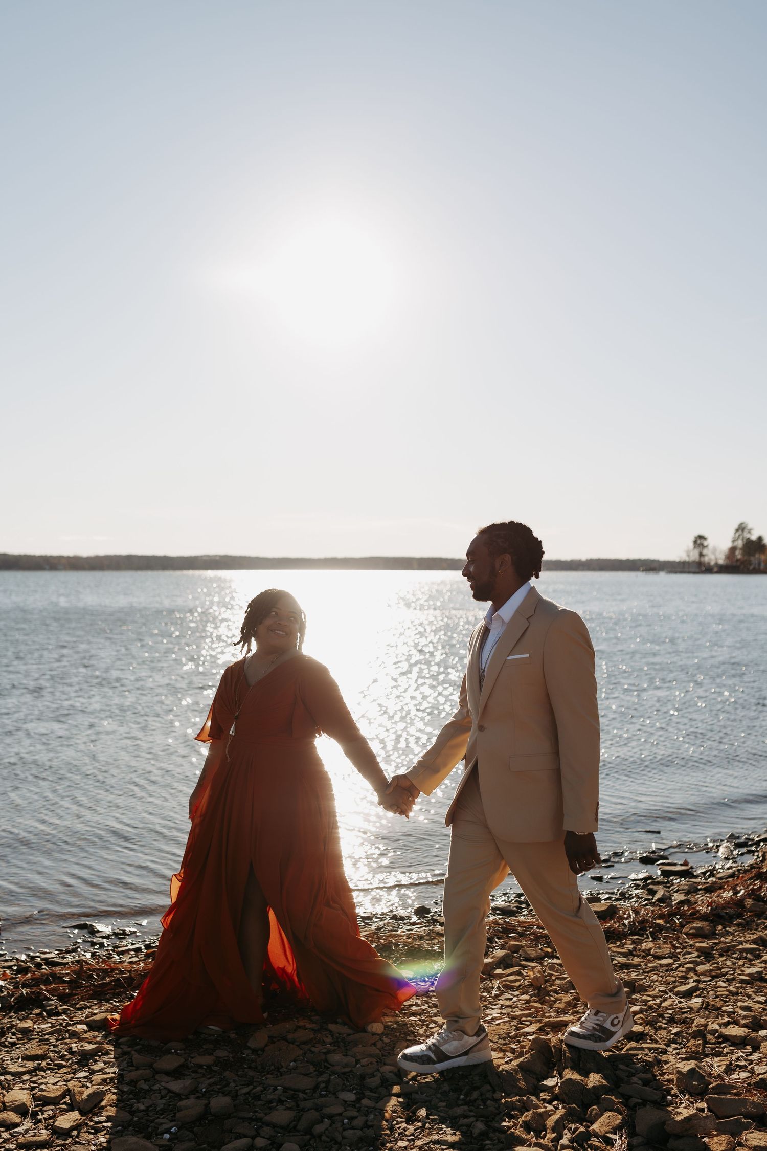 Couple holding hands by a sunlit lake, woman in red gown and man in beige suit, romantic outdoor photo.