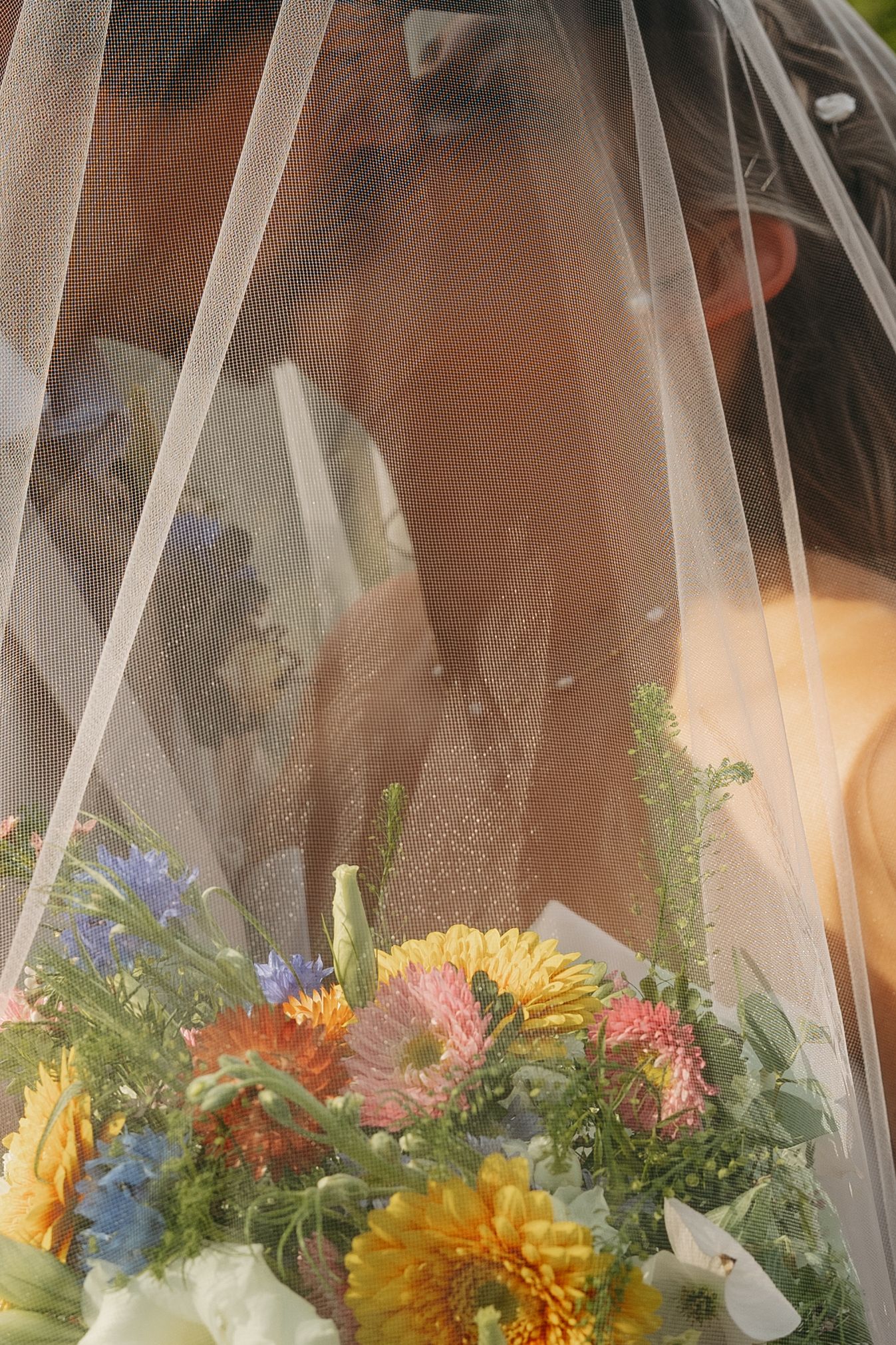 Bride and groom share a kiss behind a sheer wedding veil, with a vibrant bouquet of colorful flowers in the foreground.