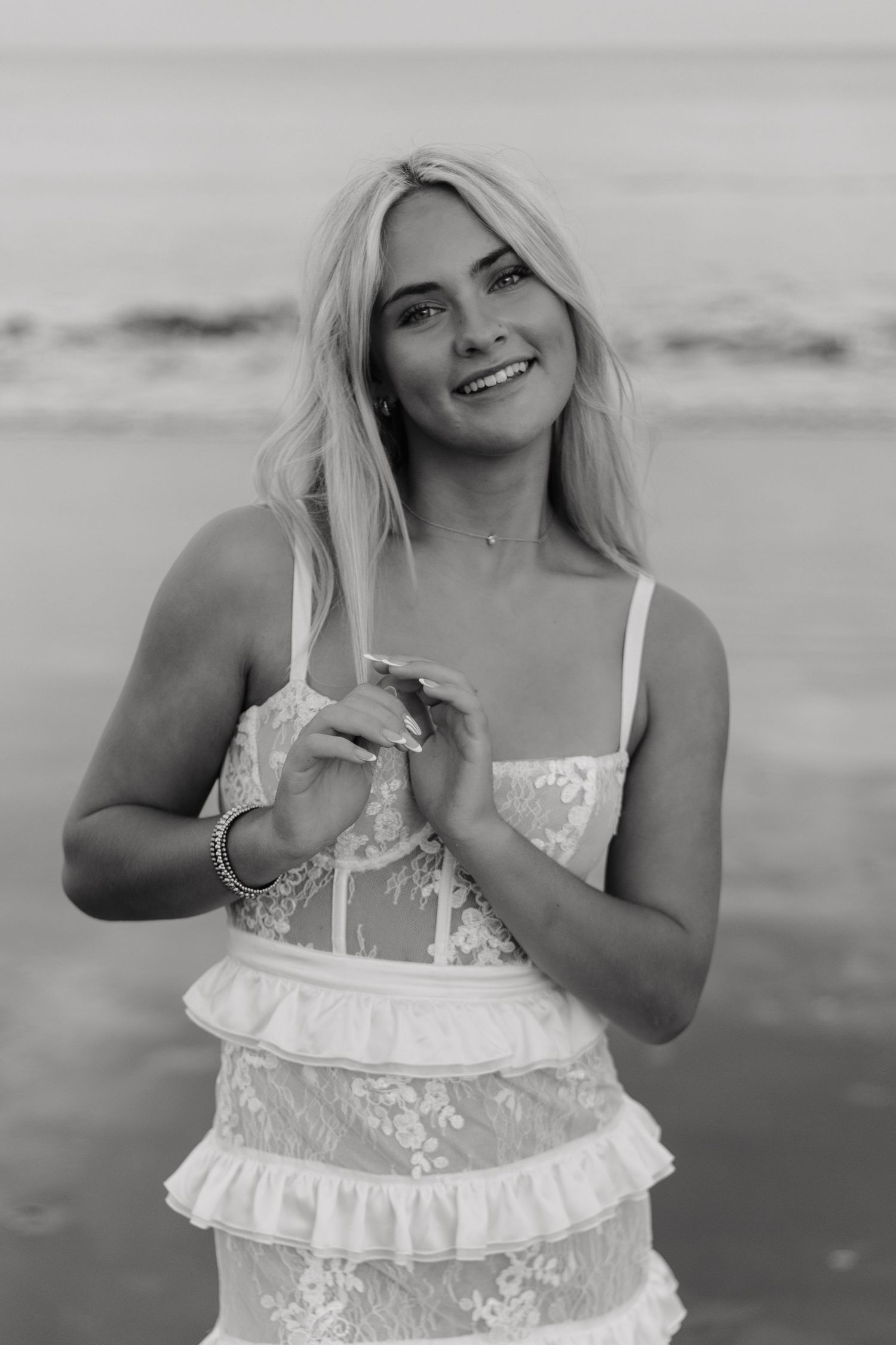 Black and white portrait of woman in lace dress with ruffles smiling at beach with ocean background.