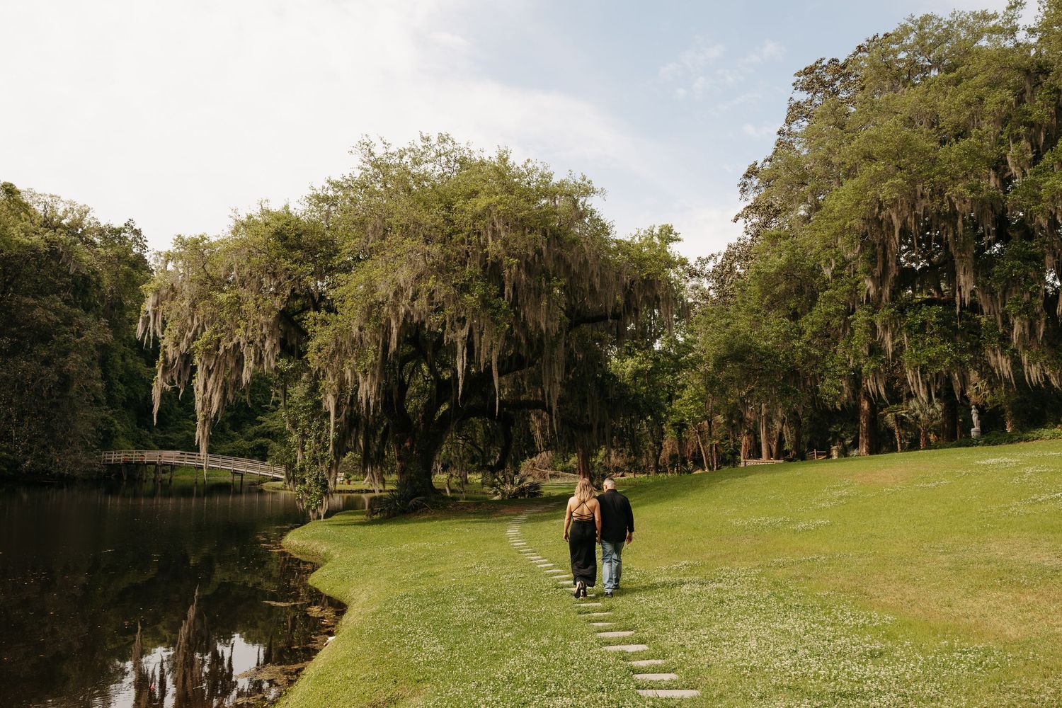 Couple walking hand in hand along a lush green lawn beside a tranquil pond beneath moss-draped oak trees.