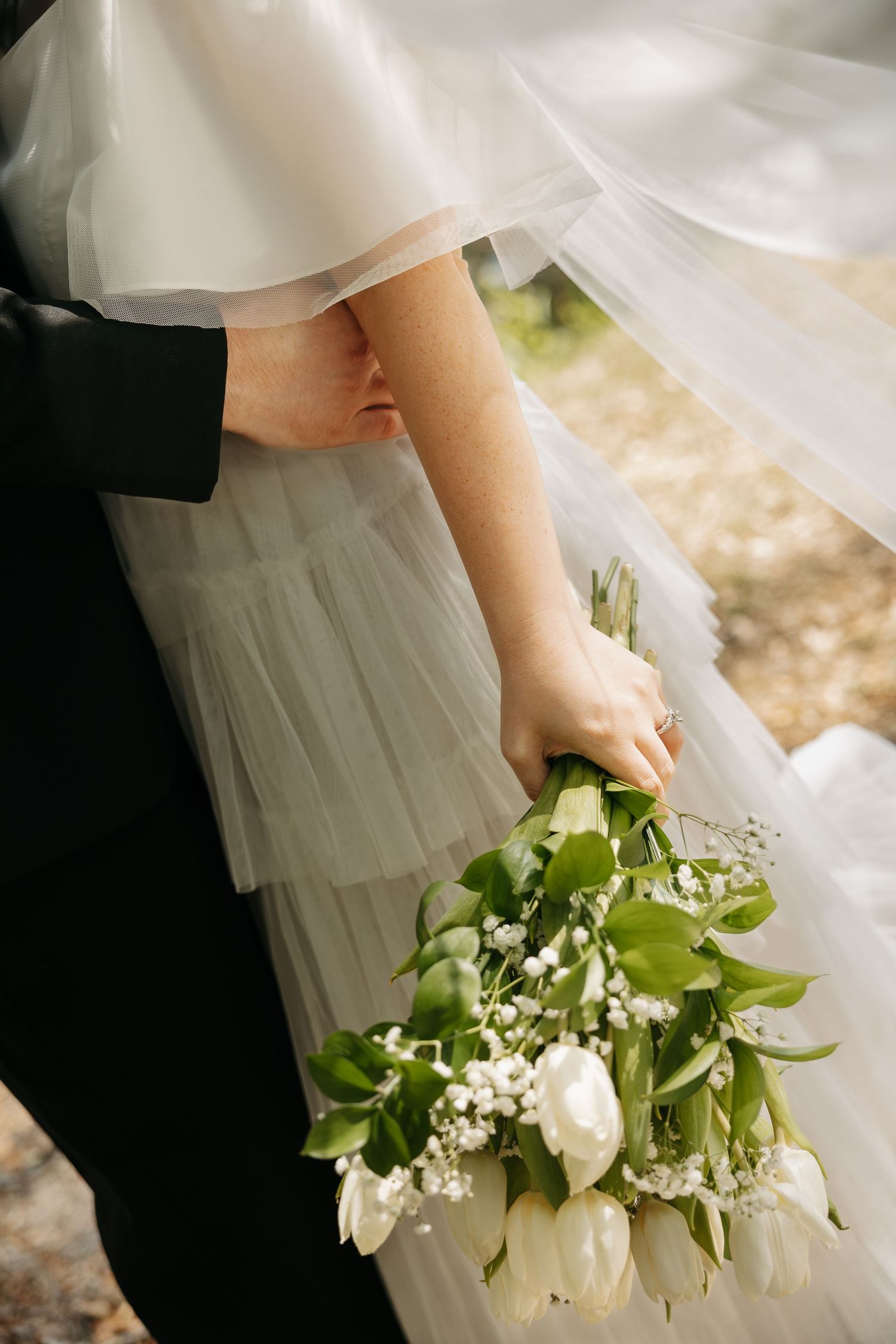 Bride holding a white tulip and baby's breath wedding bouquet while wearing a layered tulle gown beside a groom.