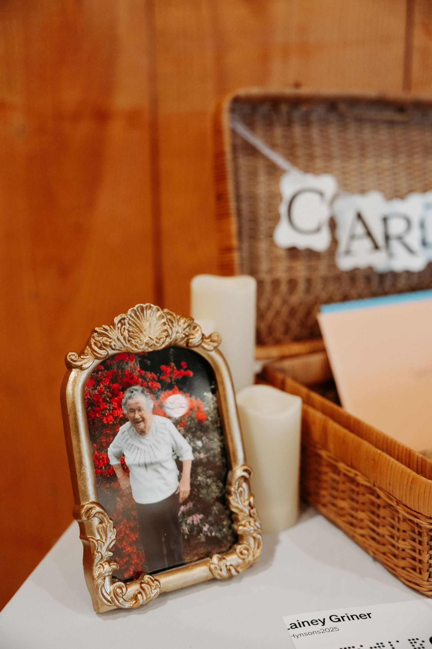 Ornate gold framed photo of elderly woman displayed on wedding card table beside wicker basket and candles.