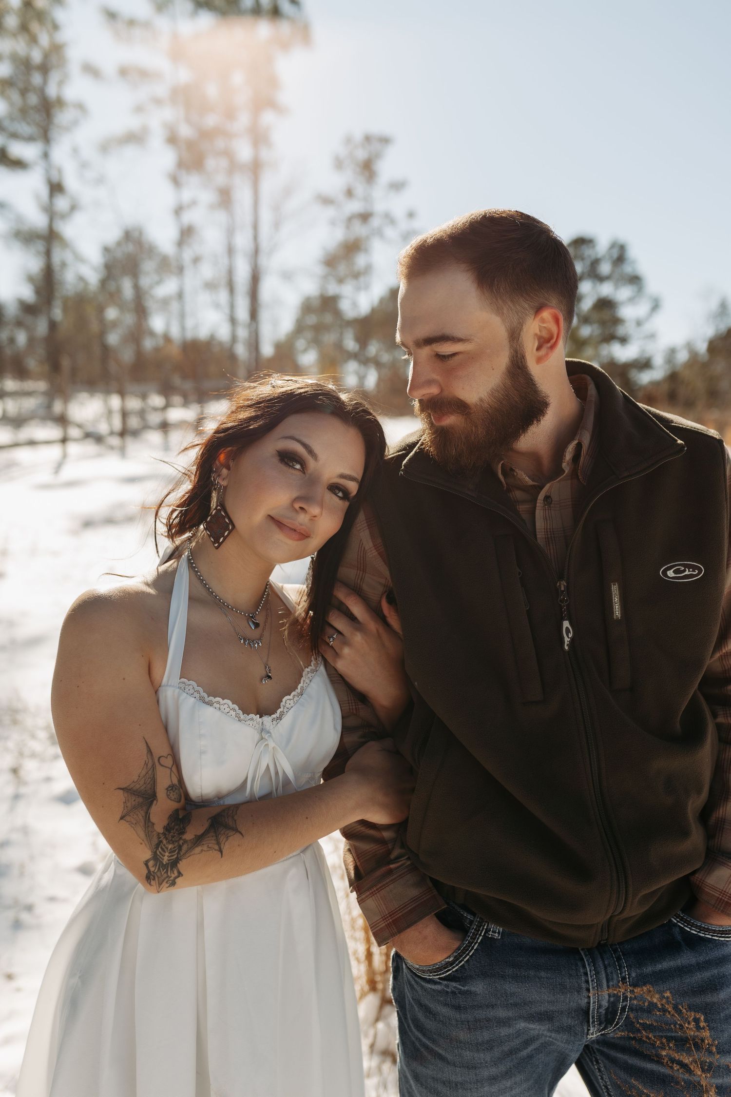 Couple embracing in snowy winter landscape, bride in white dress with tattoos, groom in brown jacket and jeans.