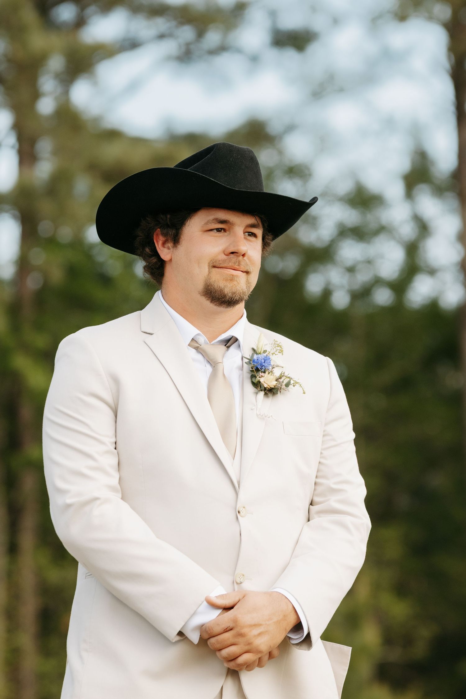 Groom in white suit and black cowboy hat with boutonniere smiling outdoors at wedding ceremony.