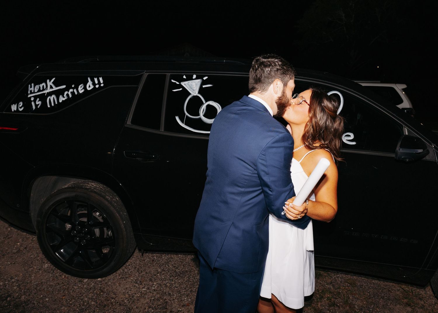 Newlyweds sharing a romantic kiss in front of a black SUV decorated with wedding messages.